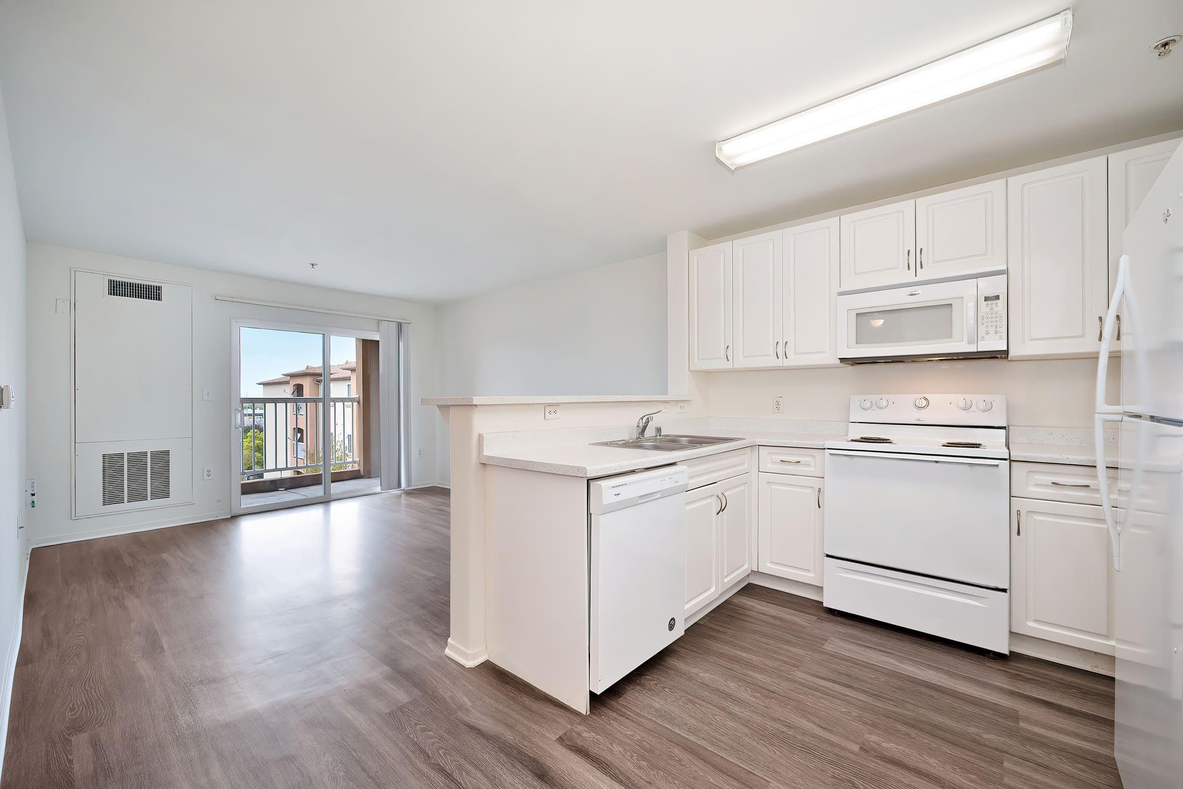 Bright and modern kitchen with white cabinetry, a microwave, and a dishwasher. The adjacent living space features large windows allowing natural light, with a view of a balcony. Light-colored walls and wood-like flooring create a spacious and inviting atmosphere.