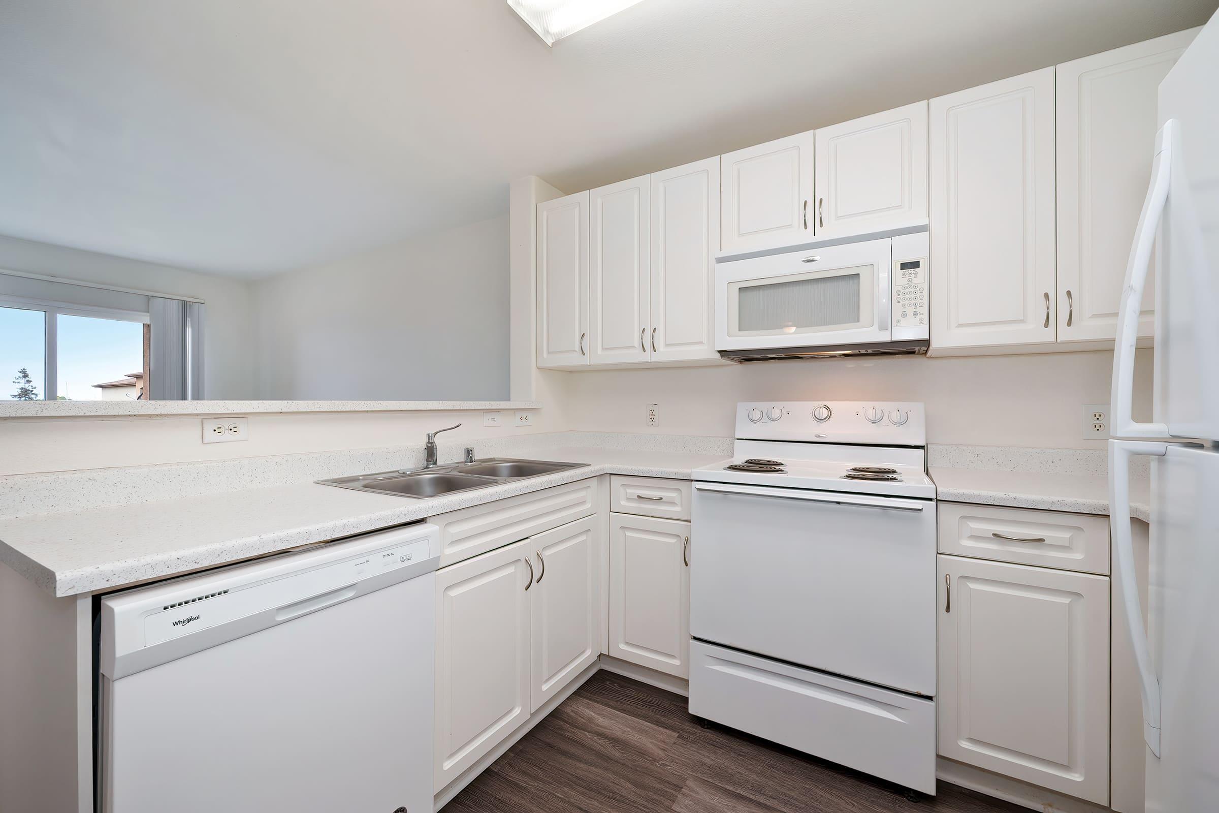 A modern kitchen featuring white cabinetry, a stainless steel double sink, a white stove and microwave, a dishwasher, and light-colored countertops. Natural light enters through a nearby window, enhancing the bright and clean aesthetic.