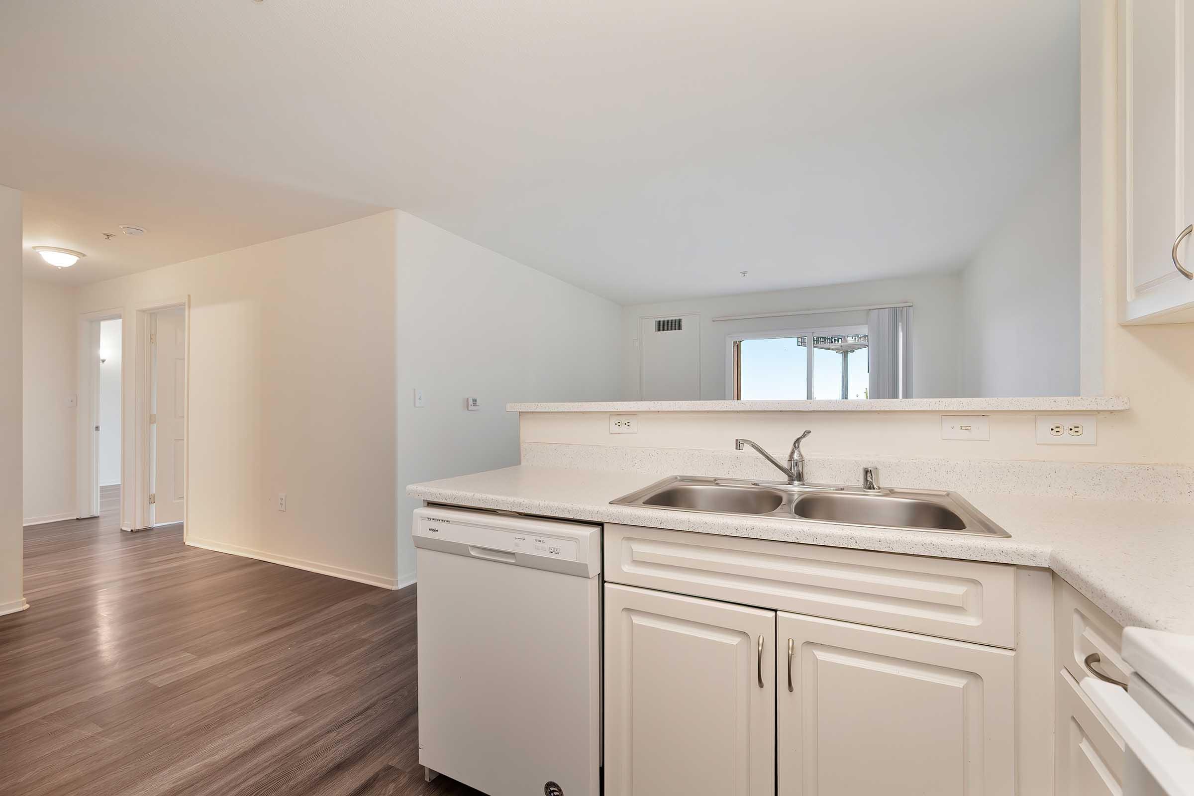 Bright and modern kitchen featuring a double sink, dishwasher, and light-colored countertops. The space is open-concept, leading into a living area with large windows and soft natural light. The flooring is a warm wood tone, enhancing the welcoming atmosphere of the room.