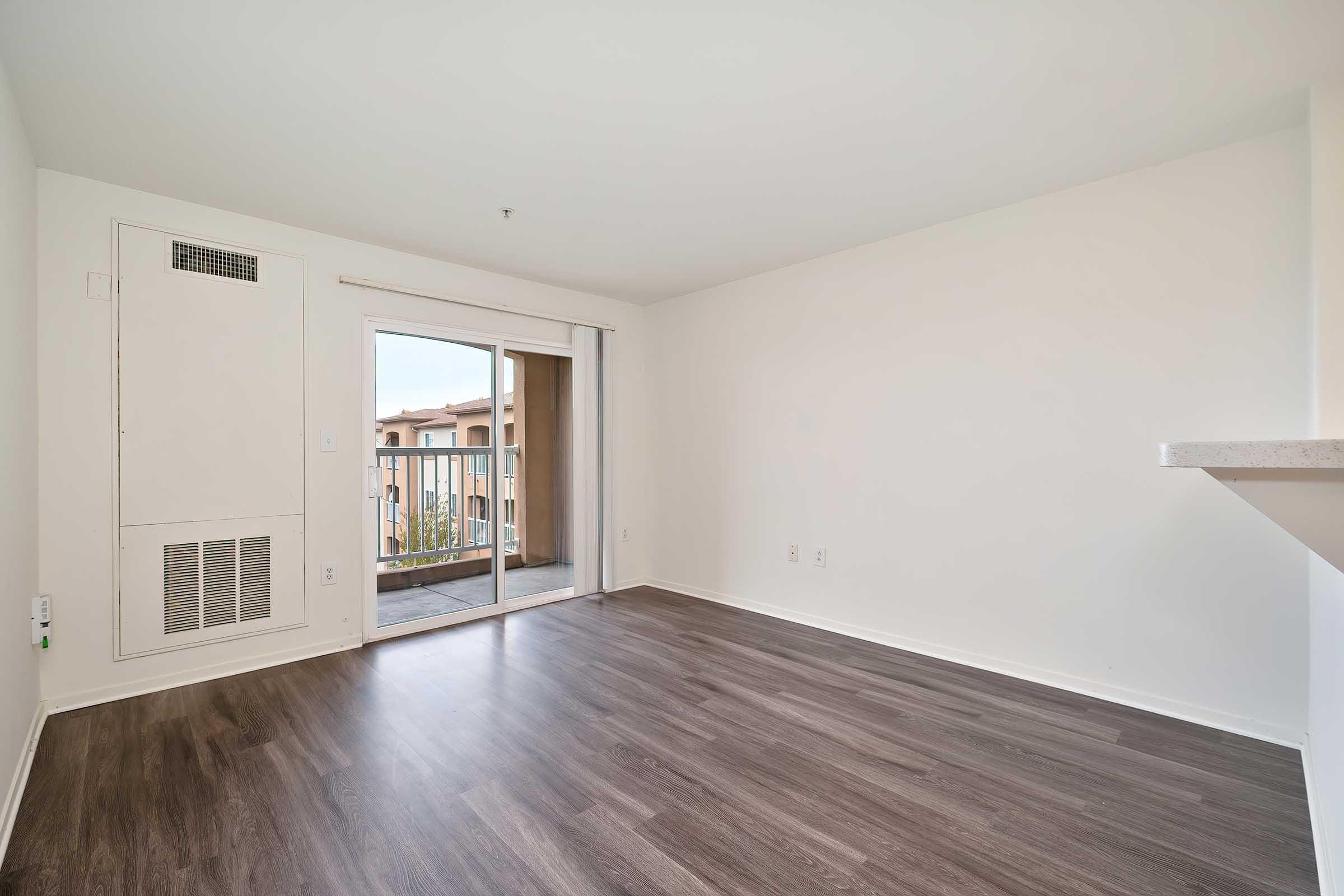 A spacious, empty room with light-colored walls and brown laminate flooring. There is a large sliding glass door leading to a balcony, providing natural light. The room features a wall-mounted vent and is free of furniture, emphasizing its open layout.