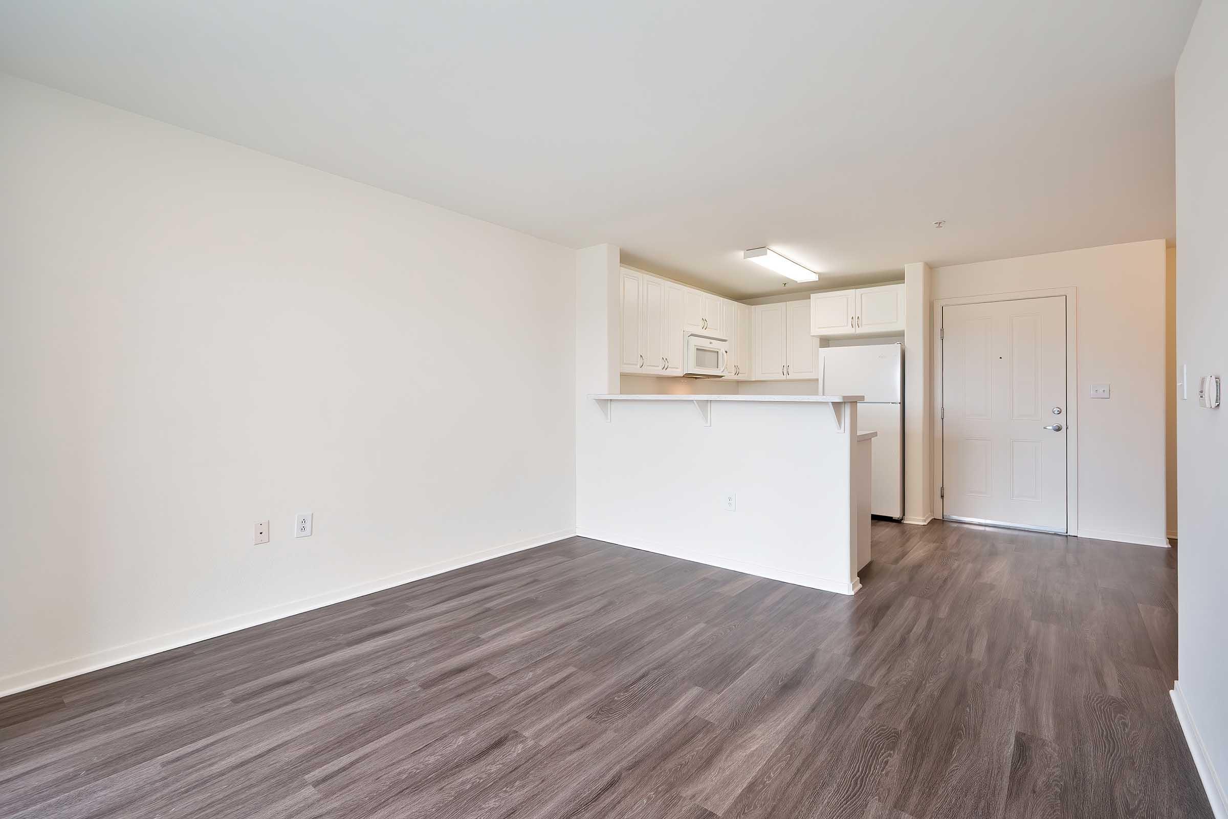 A spacious, empty room with light-colored walls and dark laminate flooring. The kitchen area features white cabinetry, a microwave, and a refrigerator, while a door leads outside. Natural light fills the space, creating a bright and inviting atmosphere.