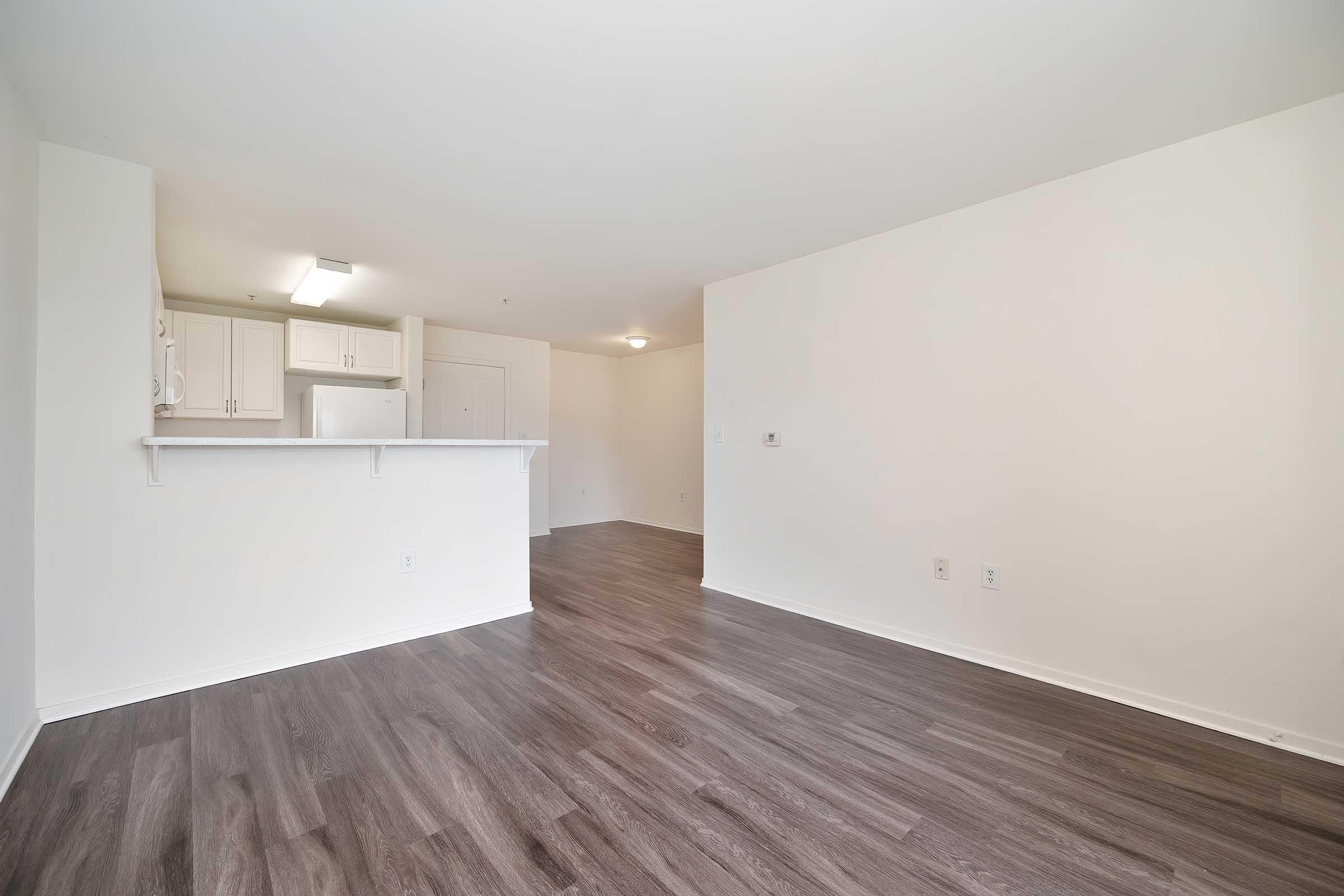 A spacious, empty room featuring light-colored walls and dark wood-like flooring. The left side includes a kitchen area with a bar counter, while the right side leads to an open area with no furniture. Natural light fills the space, creating a bright, airy atmosphere.