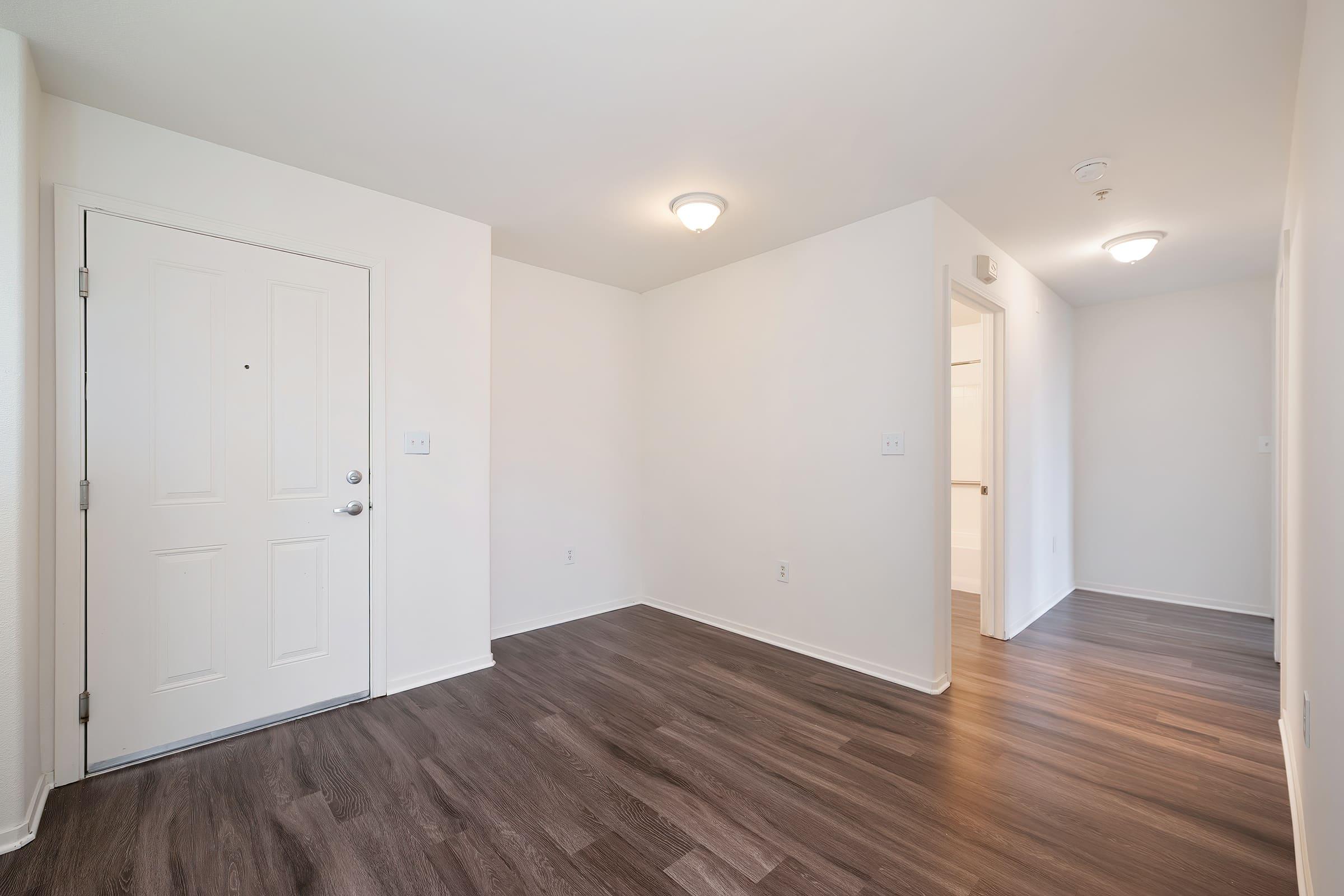 A vacant room featuring light-colored walls and dark hardwood flooring. The entrance door is on the left, providing access to a hallway leading to another room. Two ceiling lights illuminate the space, creating a bright and airy atmosphere.