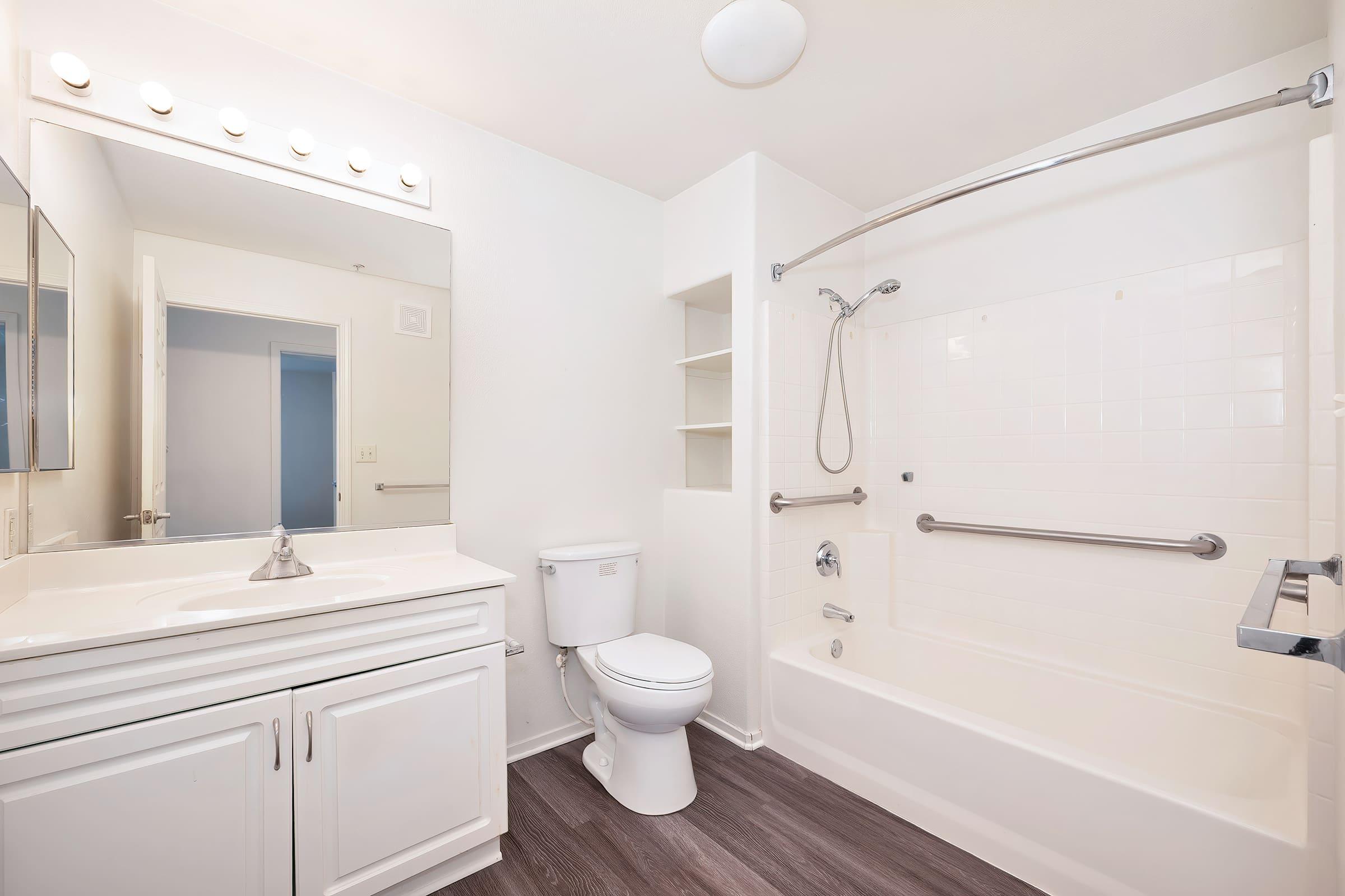 A clean and modern bathroom featuring a white bathtub with a shower, a toilet, a sink with a mirror above, and light fixtures. The walls are painted in light colors, and the floor has a wood-like finish. Shelf space is available for storage.
