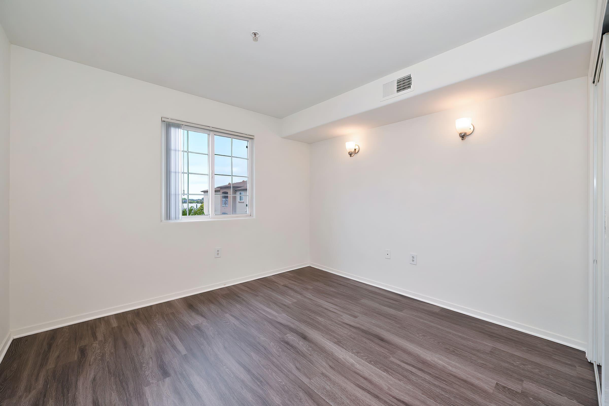 Empty room with light-colored walls and wooden flooring. There's a window on the left side allowing natural light, and two wall-mounted light fixtures. The room is spacious and minimally decorated, creating a simple and clean aesthetic.