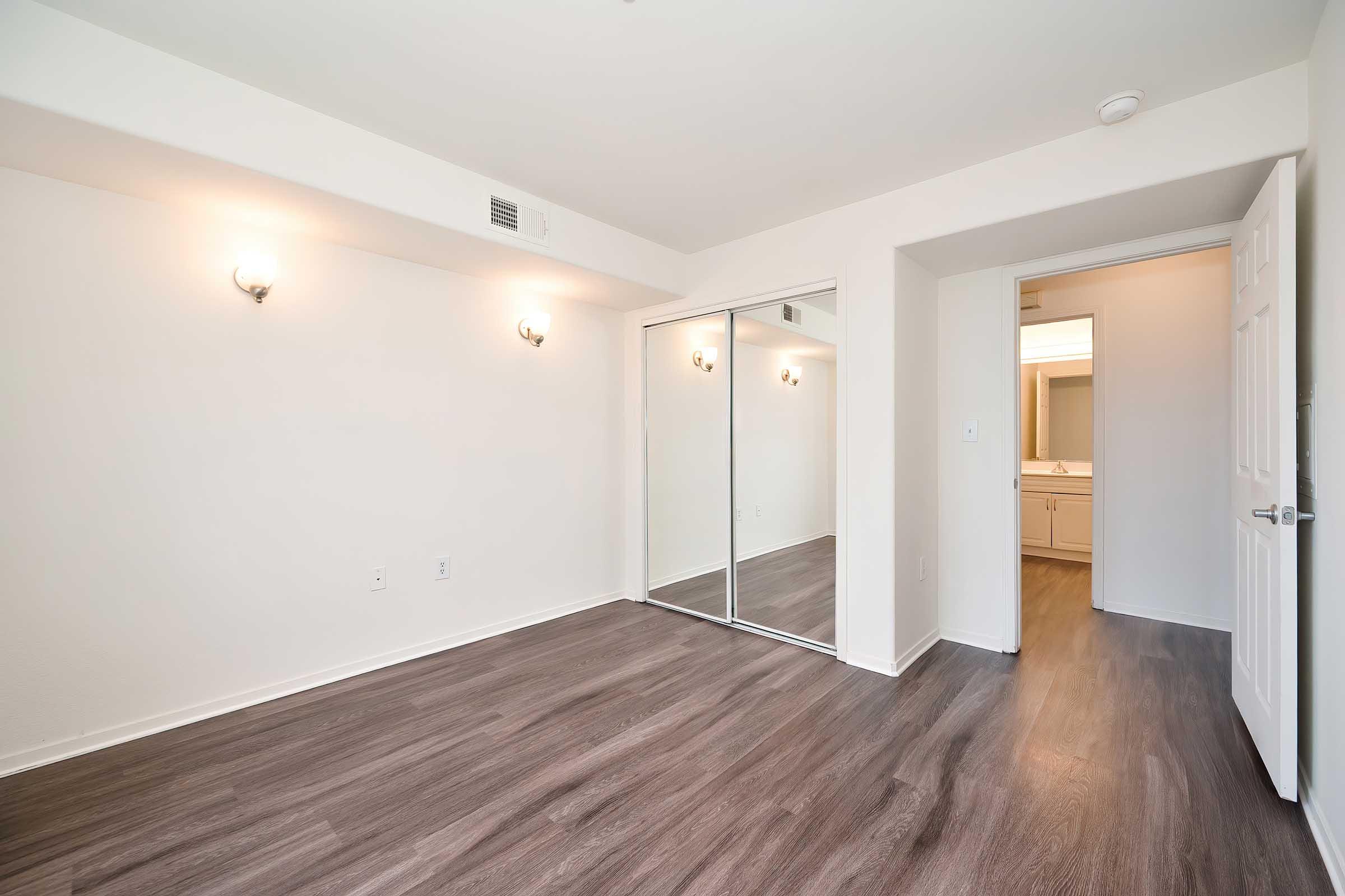 Empty room featuring light-colored walls and dark laminate flooring. It has two wall sconces for lighting and a large mirrored closet with sliding doors. A doorway on the right leads to an adjoining bathroom area with vanity lights. The overall ambiance is bright and spacious.