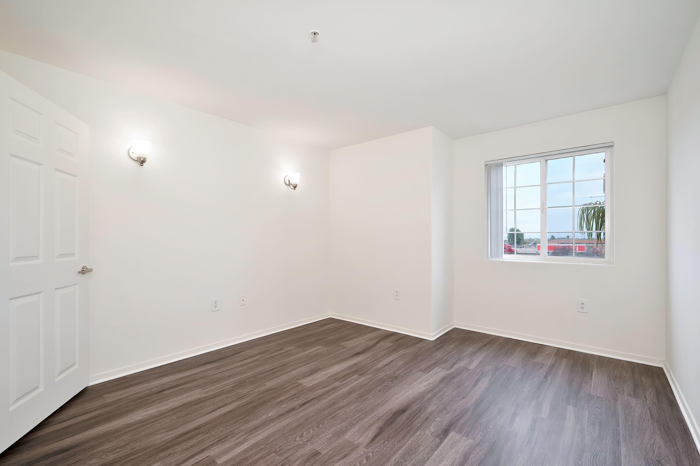 Empty room with light-colored walls, wooden flooring, and a window. Two wall sconces provide lighting, and a door is visible on one side. The room is unfurnished, creating a spacious and neutral environment suitable for various uses.