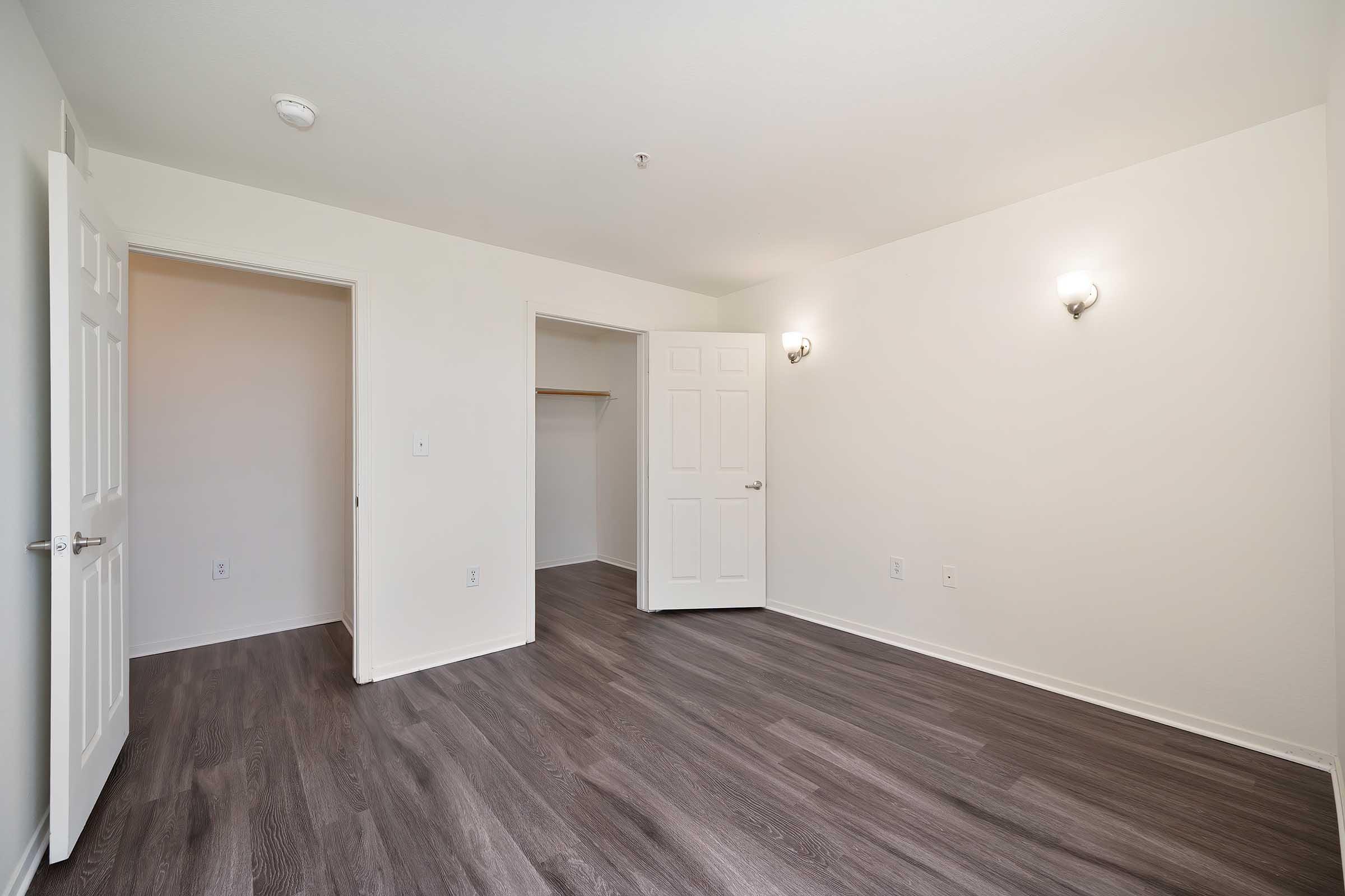 A spacious, empty room with light-colored walls and a dark wood floor. There are two doorways, one leading to a closet and the other to another room. Wall sconces provide lighting. The room is ample and ready for personal decoration or furnishings.