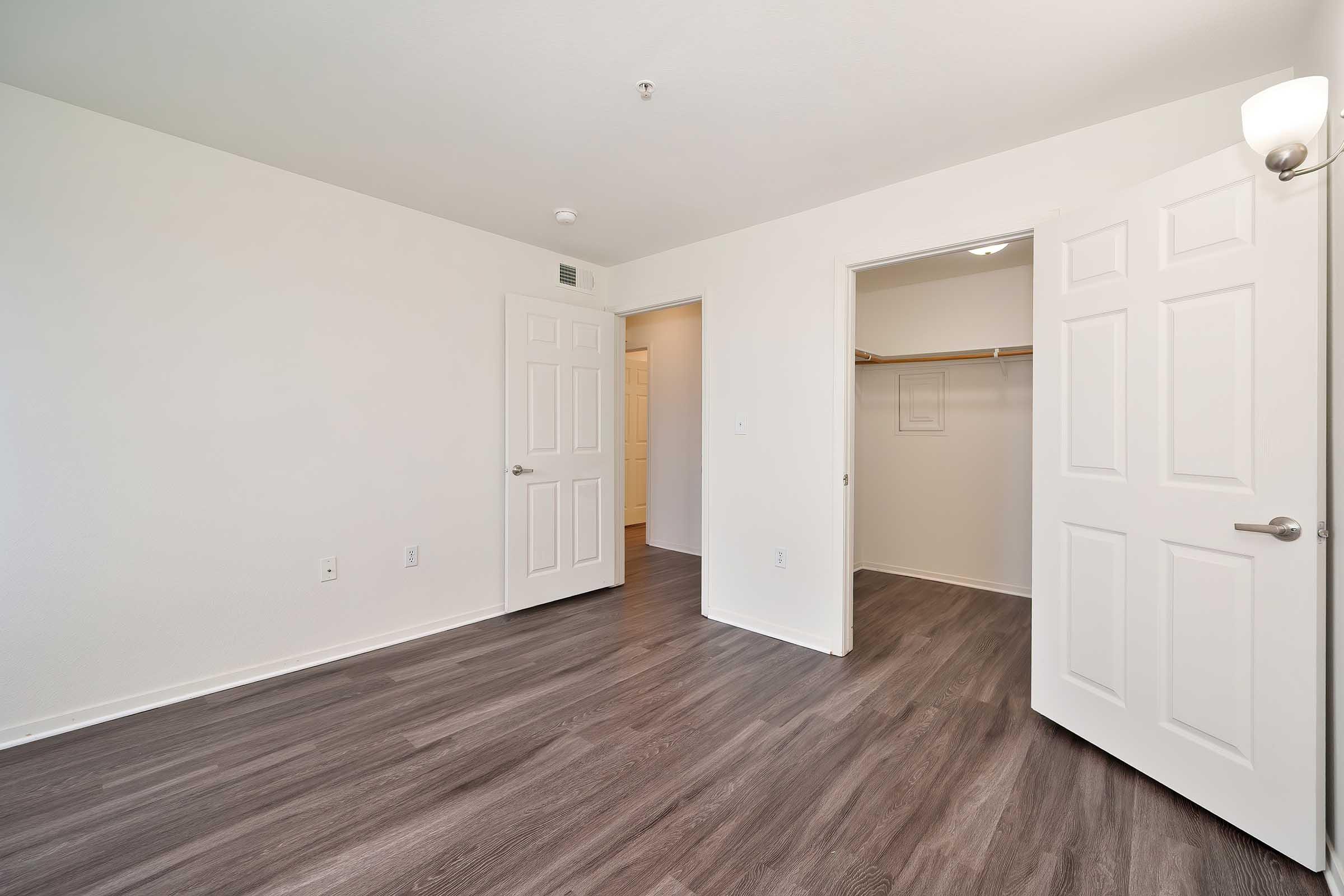 Spacious empty room with light-colored walls and dark wood-like flooring. Two open doors lead to a closet and another space. A simple light fixture is mounted on the wall, creating a bright atmosphere. The room has a clean and minimalist appearance, ready for furnishing.
