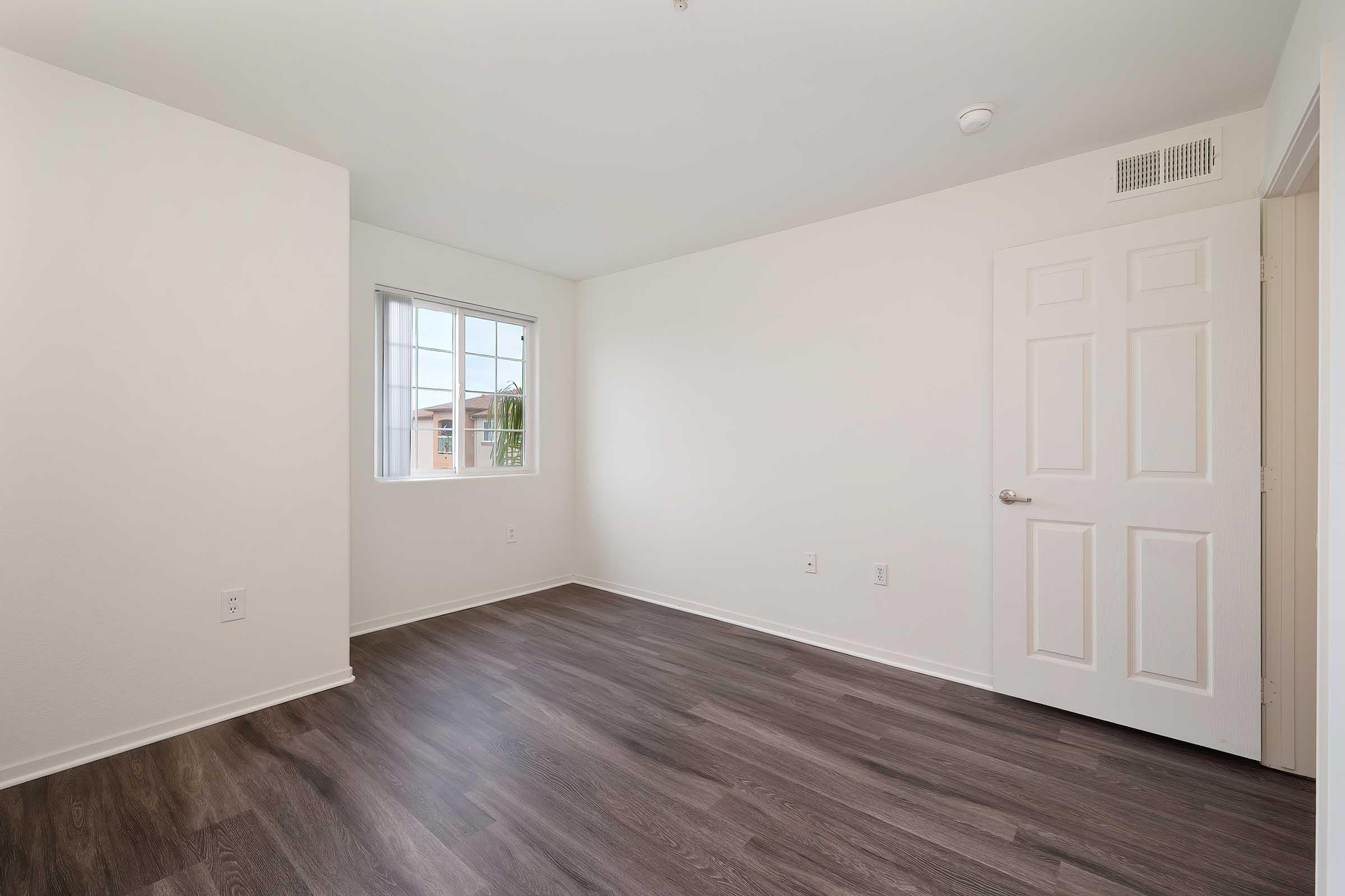 Empty room with light-colored walls and a window letting in natural light. The floor is made of dark wooden laminate. There is a closed white door on the right, and a wall-mounted light fixture is visible near the ceiling. The space is clean and uncluttered, suitable for various uses.