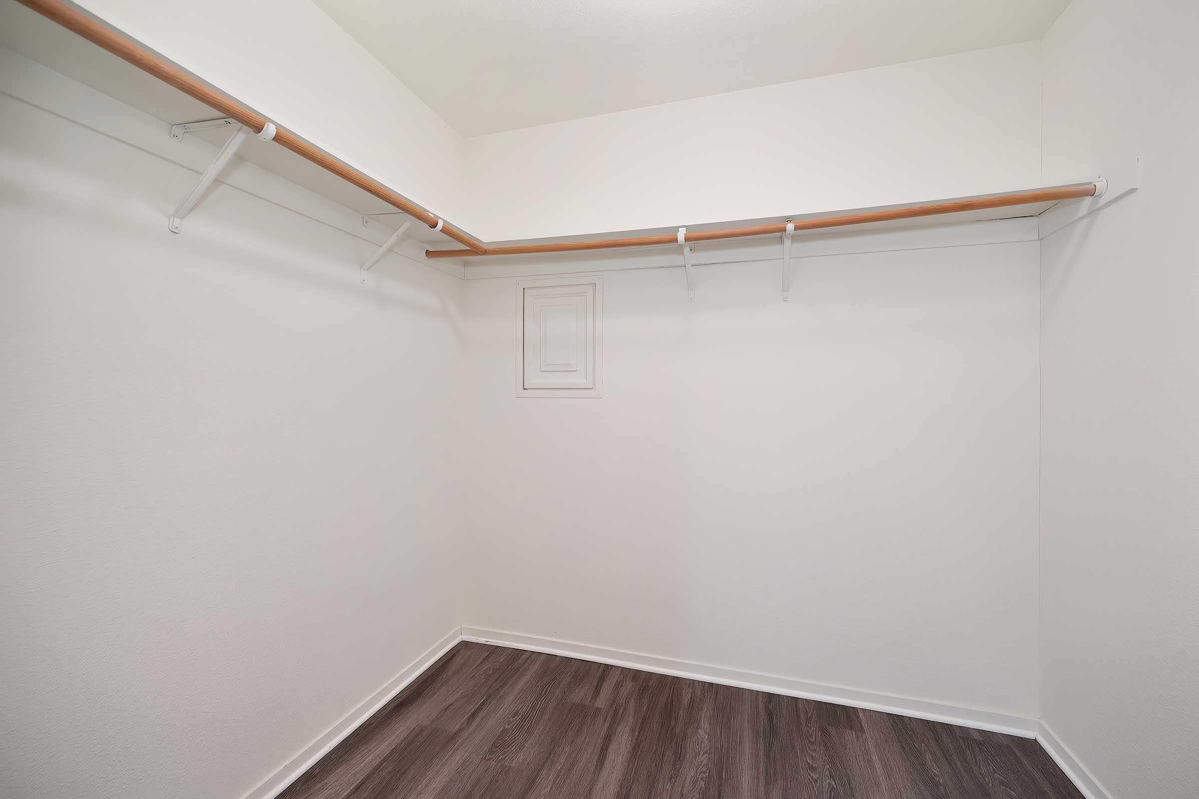 A spacious empty closet with a wooden hanging rod and white walls. There are multiple hooks along the rod for hanging clothes, and the floor is covered in dark wood laminate. A small recessed area on the wall can be seen. The overall appearance is clean and minimalistic.