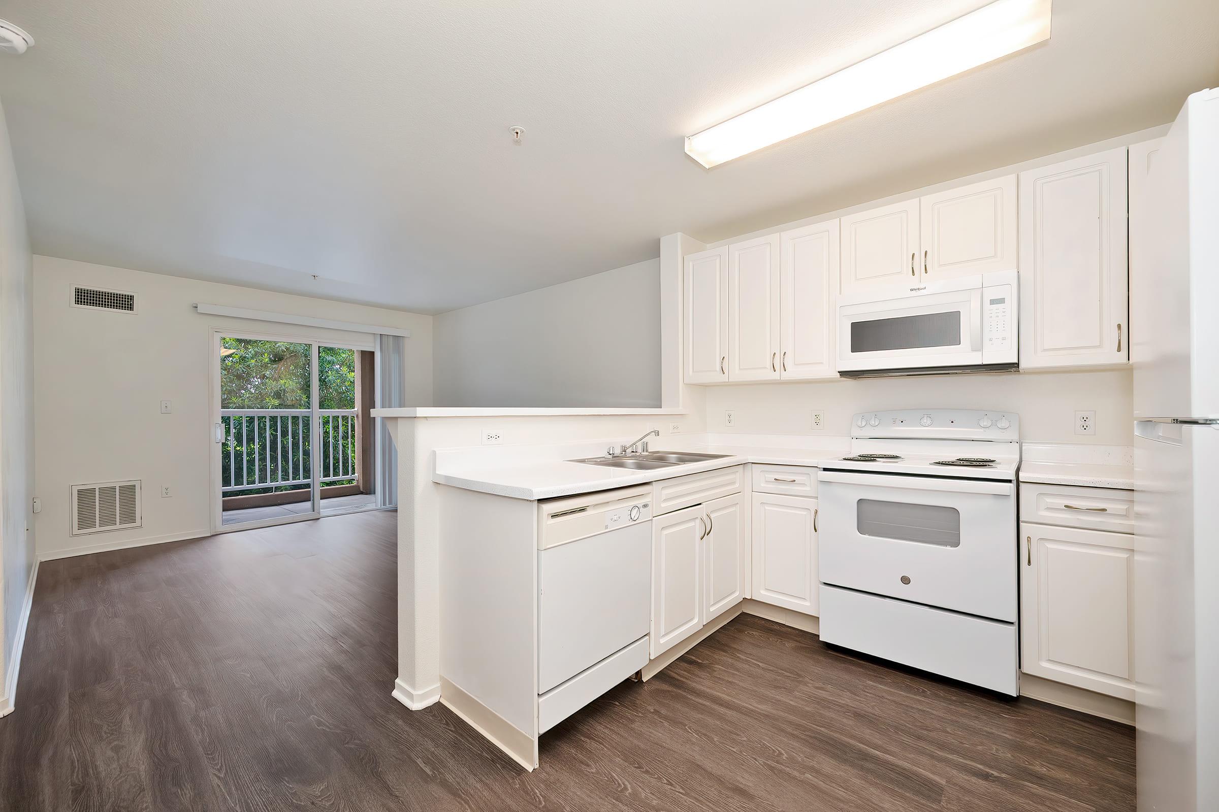 Modern kitchen featuring white cabinets, a double sink, and an oven with a microwave above. Light-colored countertops complement the clean design. A sliding glass door in the background leads to a balcony, suggesting a bright and airy living space. The floor has a wood-like finish.