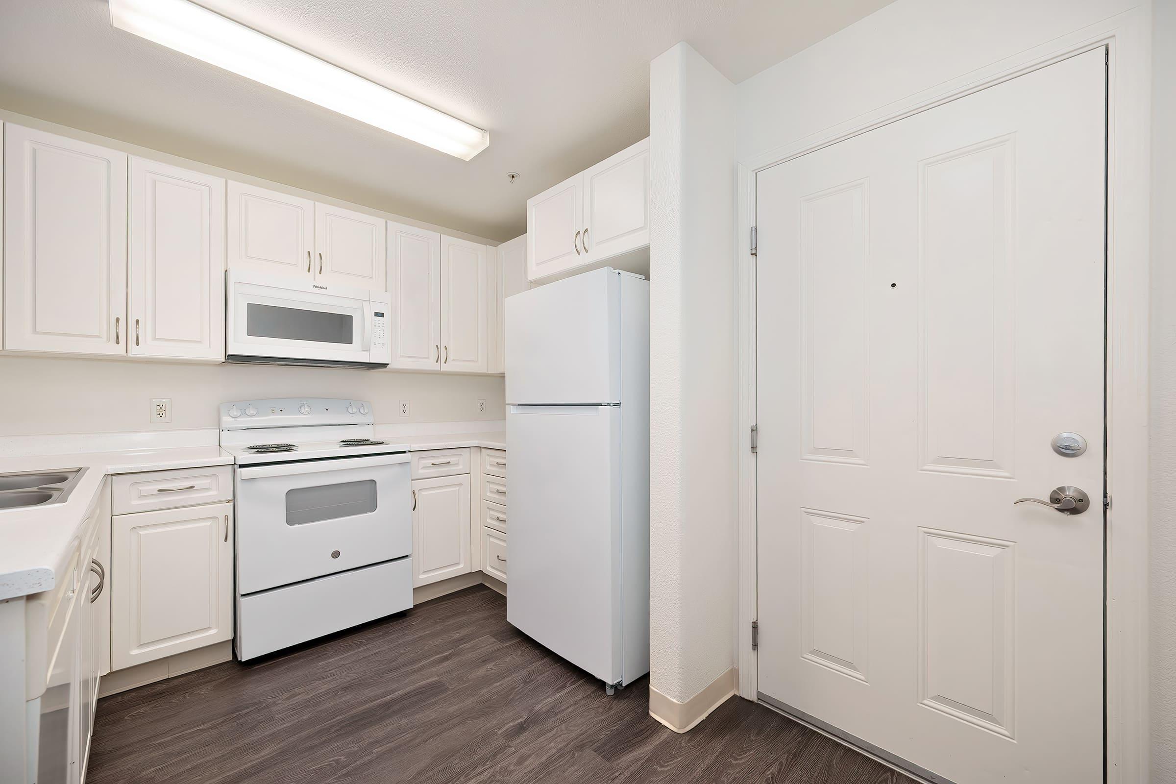A clean and modern kitchen featuring white cabinetry, a white refrigerator, a white microwave above the stove, and a light-colored countertop. The door to the kitchen is located on the right, and the flooring is dark wood. Bright overhead lighting enhances the spacious feel of the area.