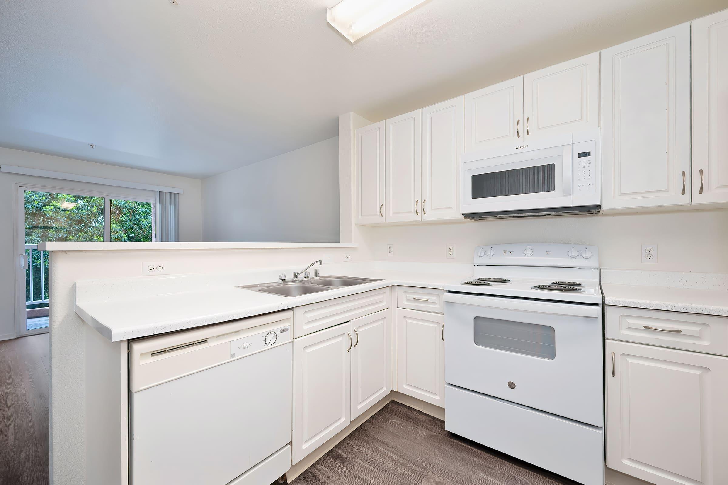 Modern kitchen featuring white cabinetry, stainless steel dishwasher, electric stove, and microwave. The countertop is light-colored with a sink, and there is a sliding door in the background leading outside. The space has a bright and airy feel, showcasing a contemporary design.