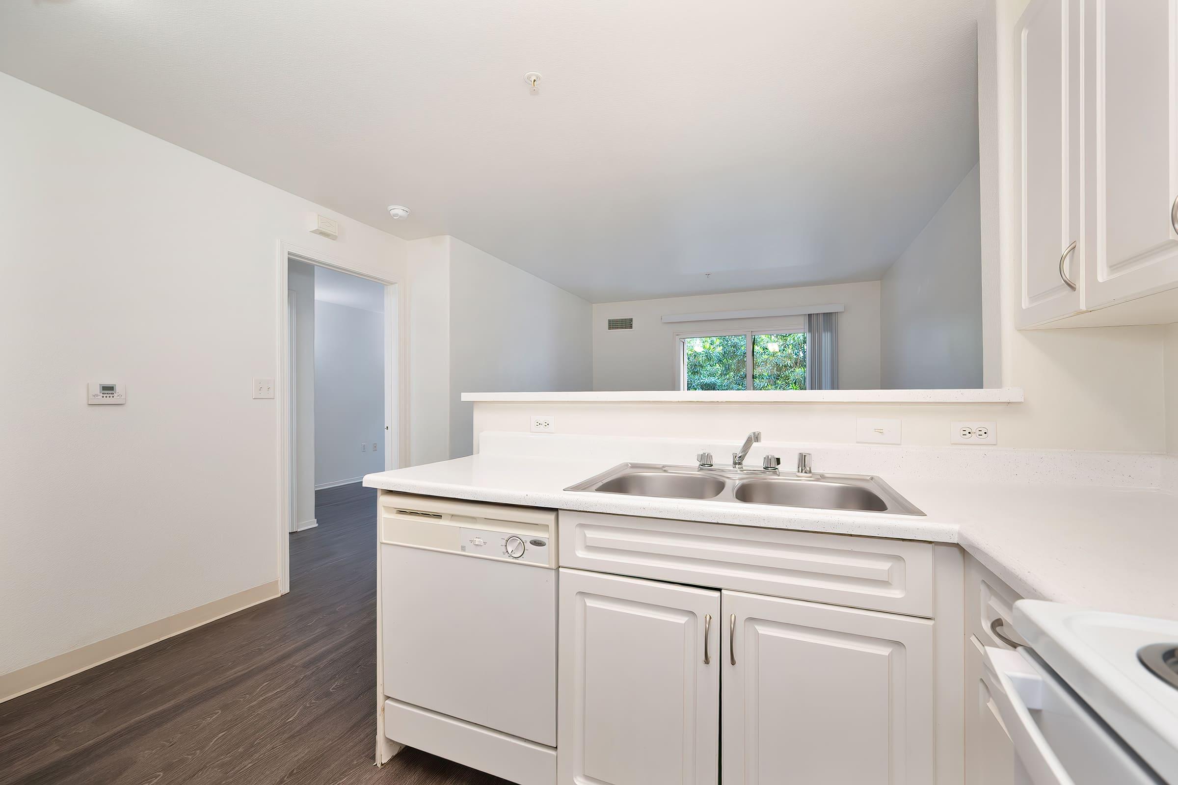 A clean, modern kitchen with white cabinets and a double sink, featuring a dishwasher nearby. The room has light-colored walls and a window visible in the background. The flooring is dark wood, providing a nice contrast to the cabinetry. The space appears bright and inviting, with natural light coming in from the window.