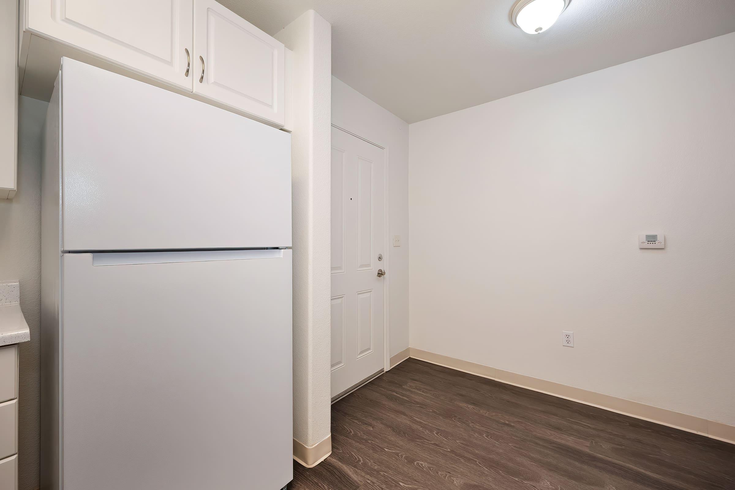 A clean and minimalistic kitchen corner featuring a white refrigerator beside a door. The walls are painted in a light color, and the floor has a dark wood-like finish. A light fixture is visible on the ceiling, adding brightness to the space.
