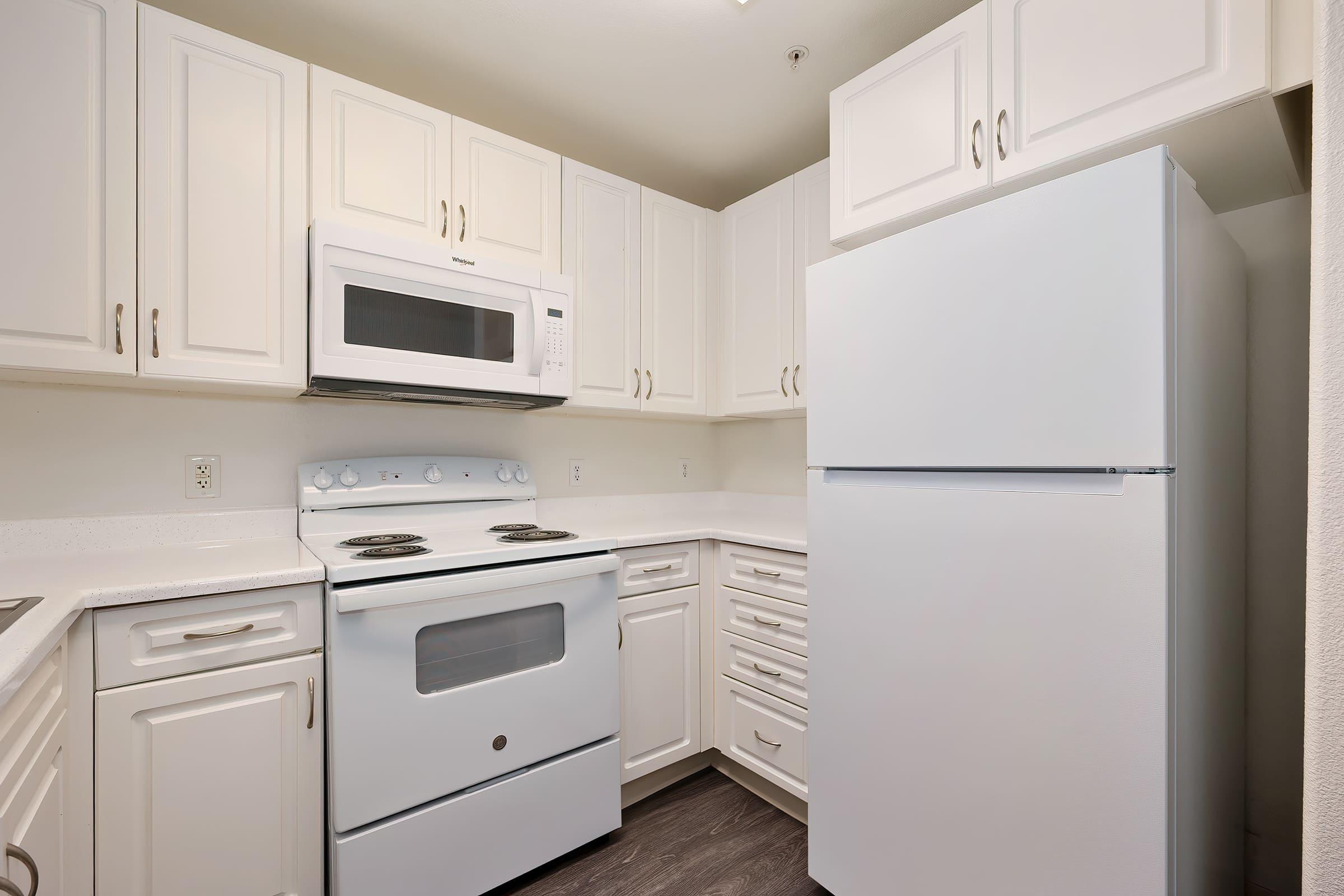 A compact kitchen featuring white cabinetry, a white refrigerator, a built-in microwave above the stovetop, and an electric oven. The countertops are light-colored, and the flooring is dark, creating a modern look. The layout is efficient, maximizing space in a small area.