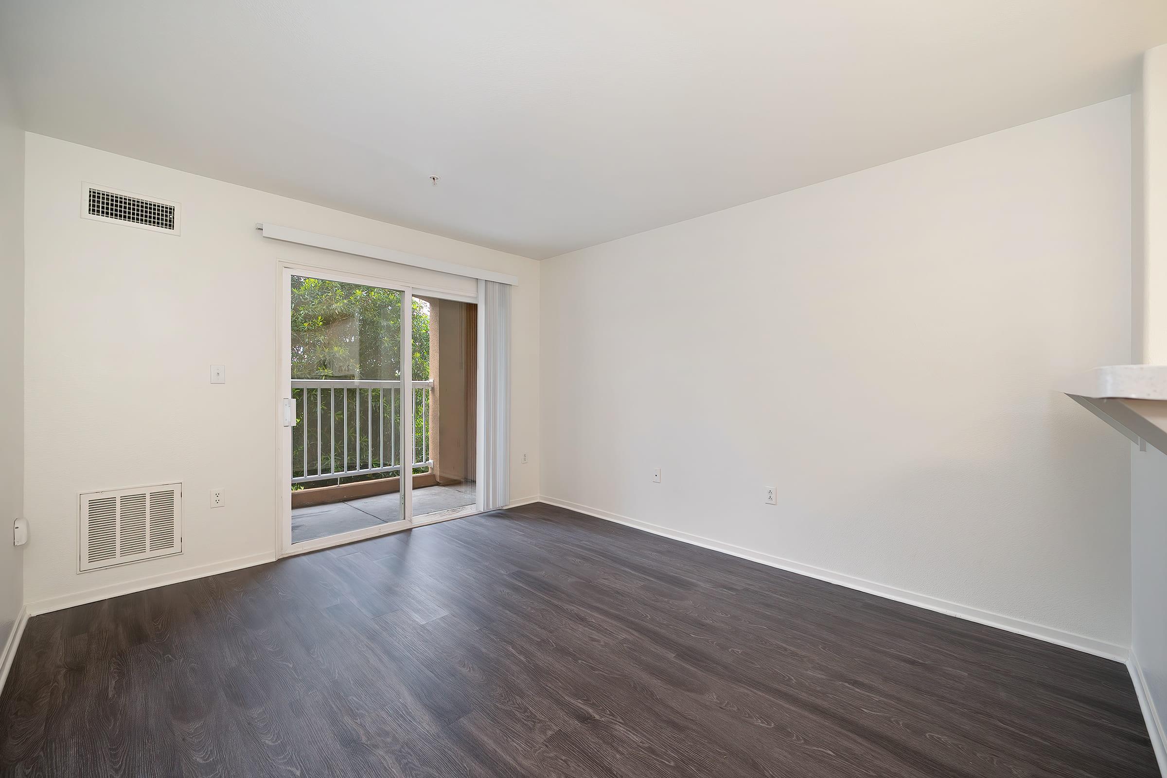 Empty living room with a light-colored wall and dark wood flooring. One wall features a sliding glass door leading to a balcony. The room is well-lit with natural light, and there is a vent on one wall. The overall space appears clean and ready for furnishing.