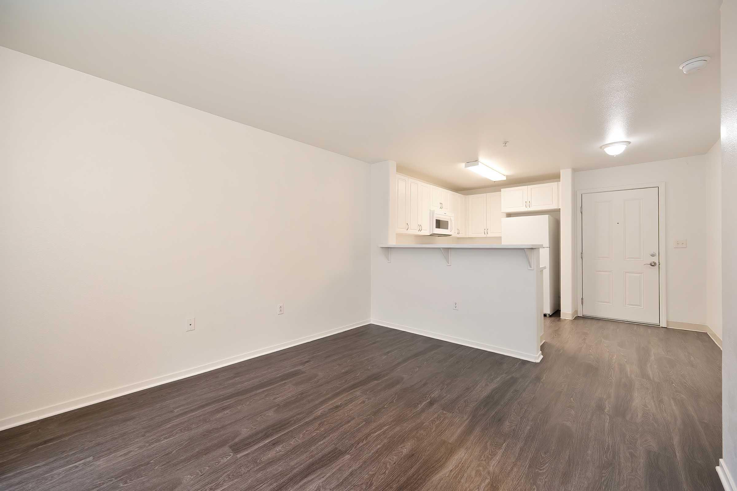 A spacious, empty room featuring light-colored walls and dark hardwood flooring. The kitchen area is visible at the far end, equipped with modern white cabinets, a microwave, and a refrigerator. A door to the right leads to the entrance, while natural light illuminates the space.