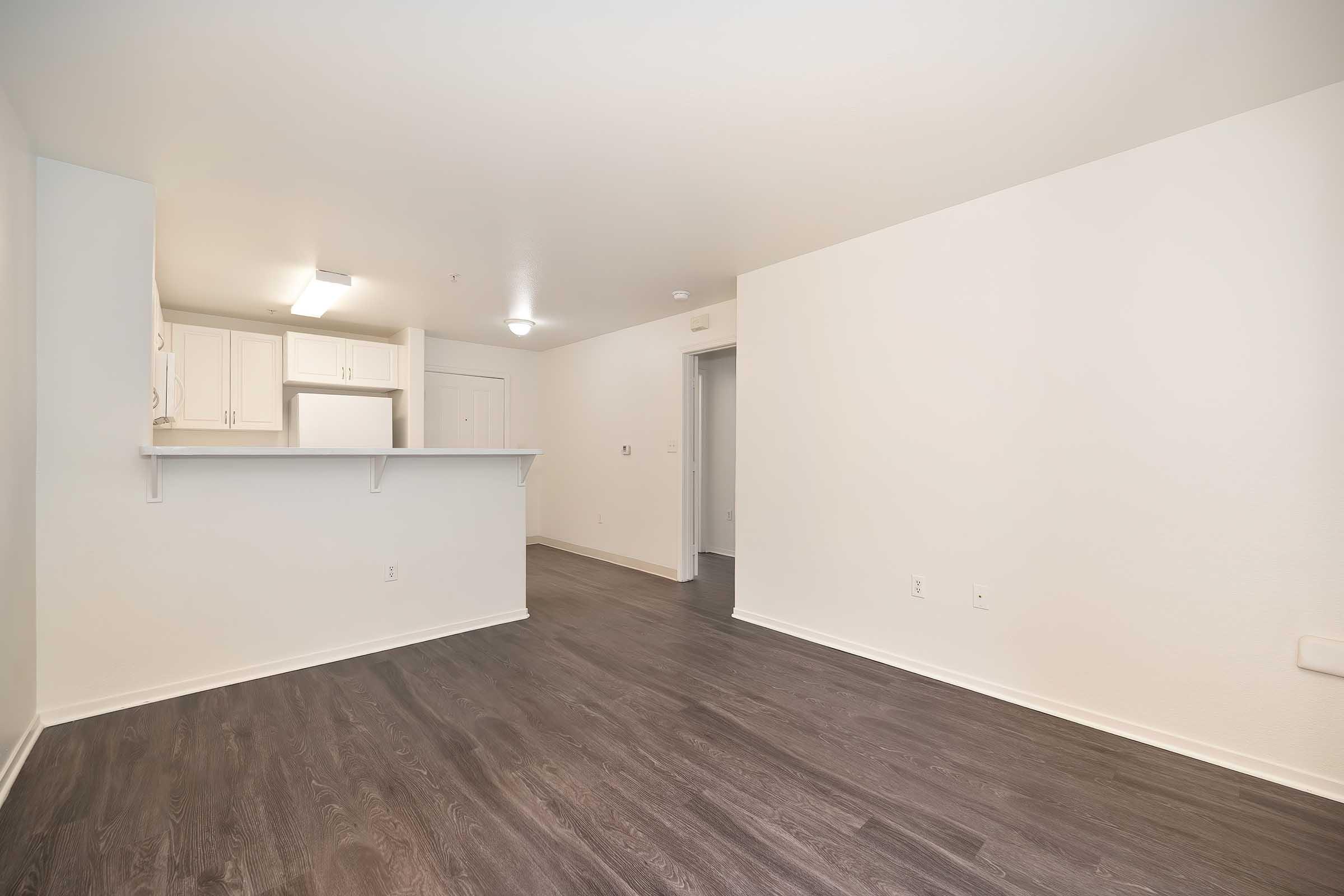A spacious, empty room with light-colored walls and dark hardwood flooring. The room features a kitchenette area with white cabinets and bright overhead lights. There is a doorway leading to another room and a clean, minimalist aesthetic.