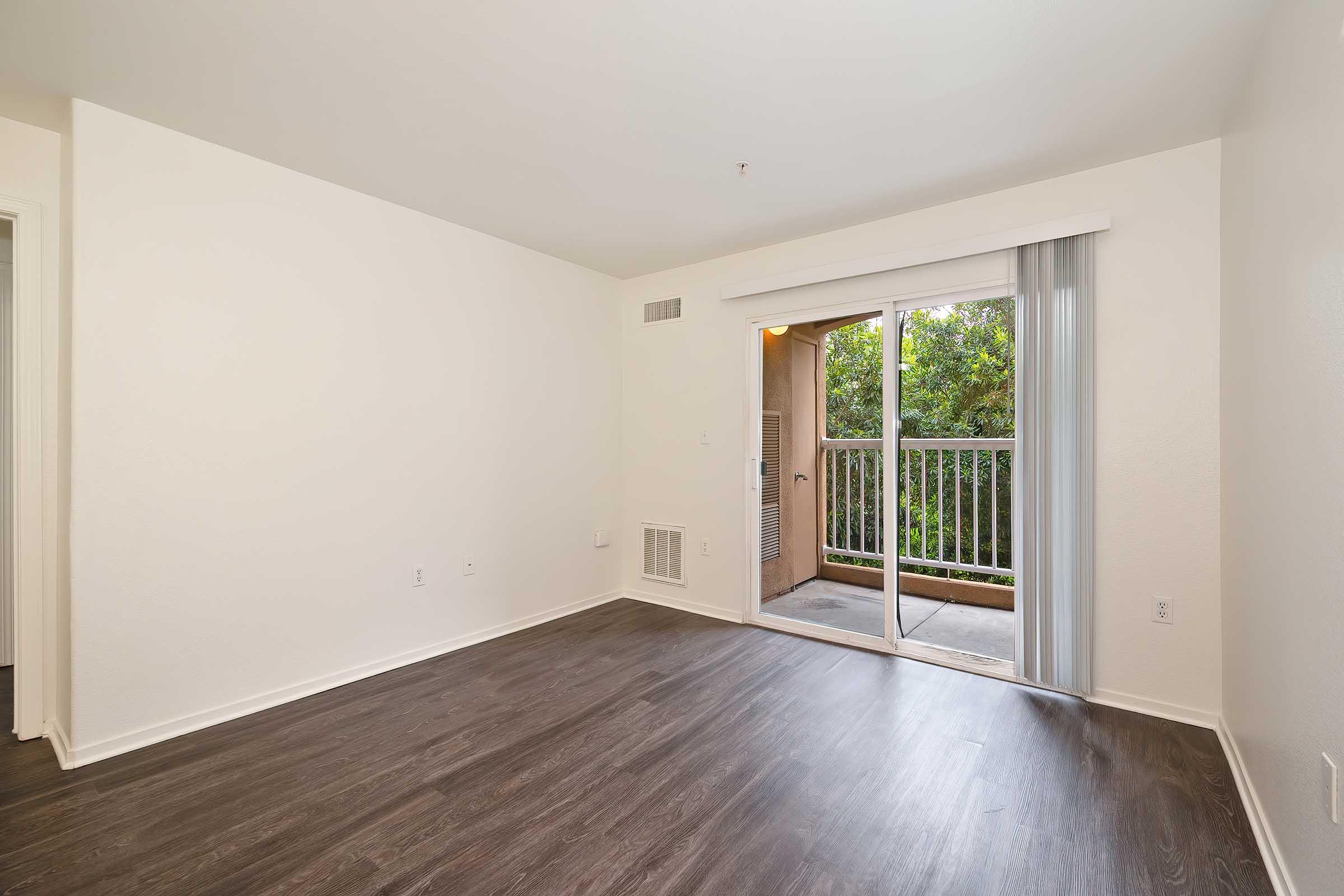 Empty room with light-colored walls and dark hardwood flooring. A sliding glass door opens to a small balcony with greenery visible outside. The room appears spacious and well-lit, with a vent on the wall and a light fixture on the ceiling.