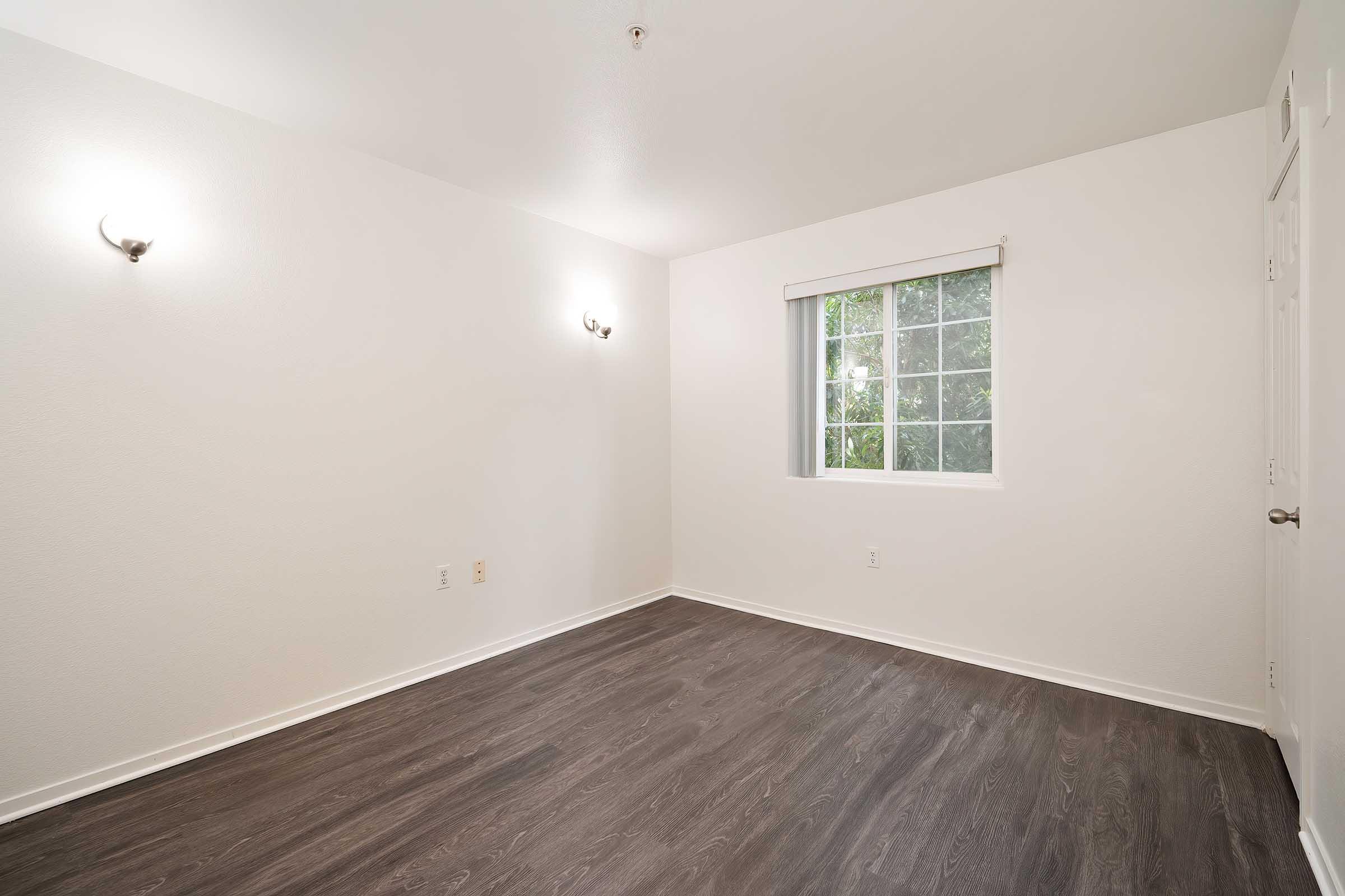 Empty room featuring light-colored walls, two wall sconces providing soft lighting, and a single window with white blinds. The floor is dark wood, and the space is free of furniture, creating a minimalist and open environment.