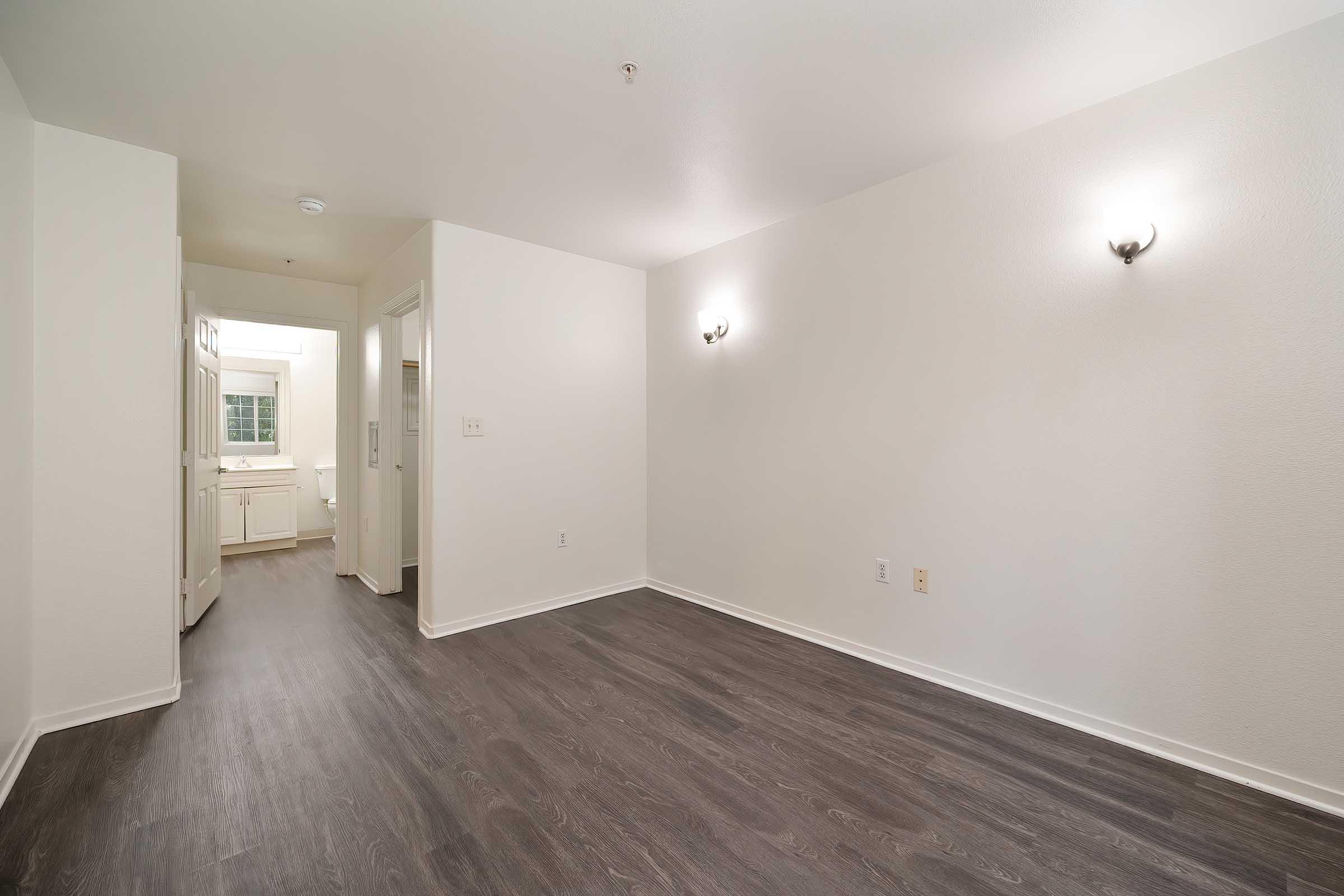 A spacious, empty room with light-colored walls and dark wooden flooring. Two wall-mounted light fixtures illuminate the space. To the left, a doorway leads to a kitchen area, while a closet can be seen in the background. The overall ambiance is bright and clean, ideal for personalizing with furniture.