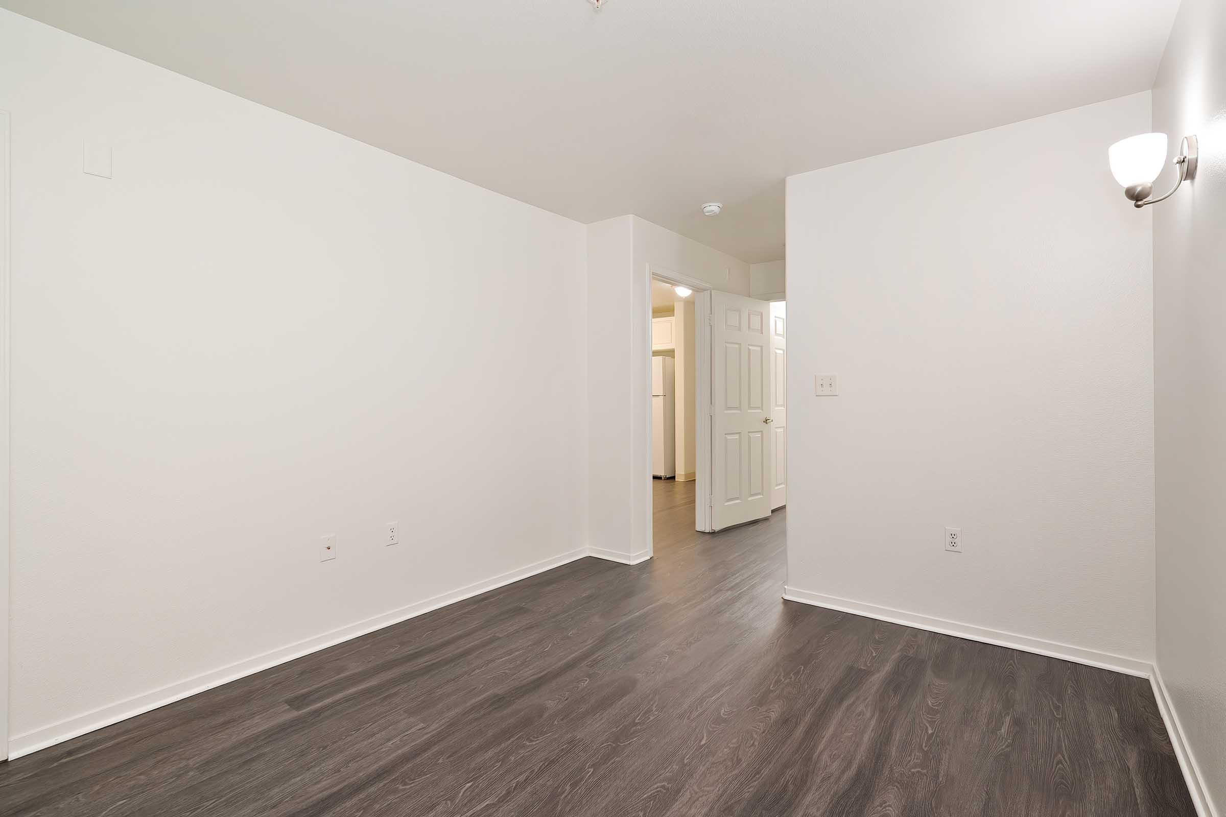 Empty room with light-colored walls and a dark wood-style floor. A wall-mounted light fixture is visible. The room has one doorway leading to another area, suggesting a spacious layout. The overall ambiance is clean and minimalistic, ideal for various uses or personal decoration.