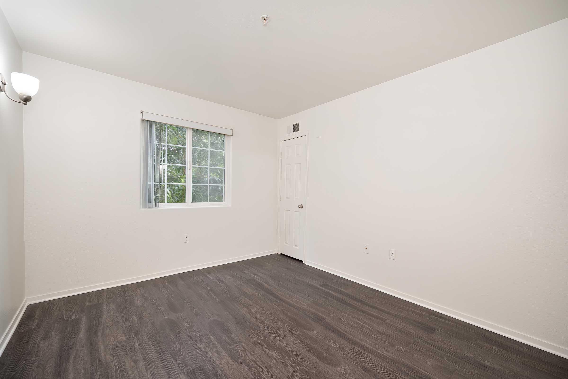A clean, empty room with light-colored walls and a window. The floor is dark wood, and there is a light fixture on the wall. The door is closed, and the overall atmosphere is bright and spacious.
