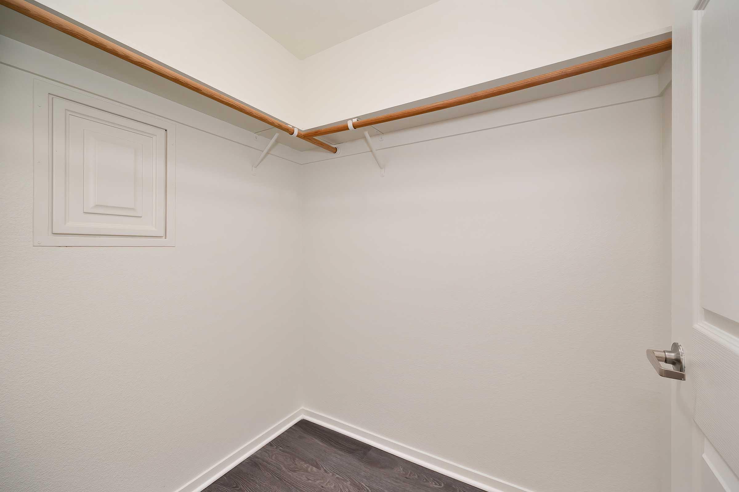 Empty closet with a single wooden clothes rod and two metal hooks mounted on the wall. The walls are painted white, and the floor features dark laminate flooring. There is a small panel on one wall next to the rod. The overall appearance is clean and uncluttered.