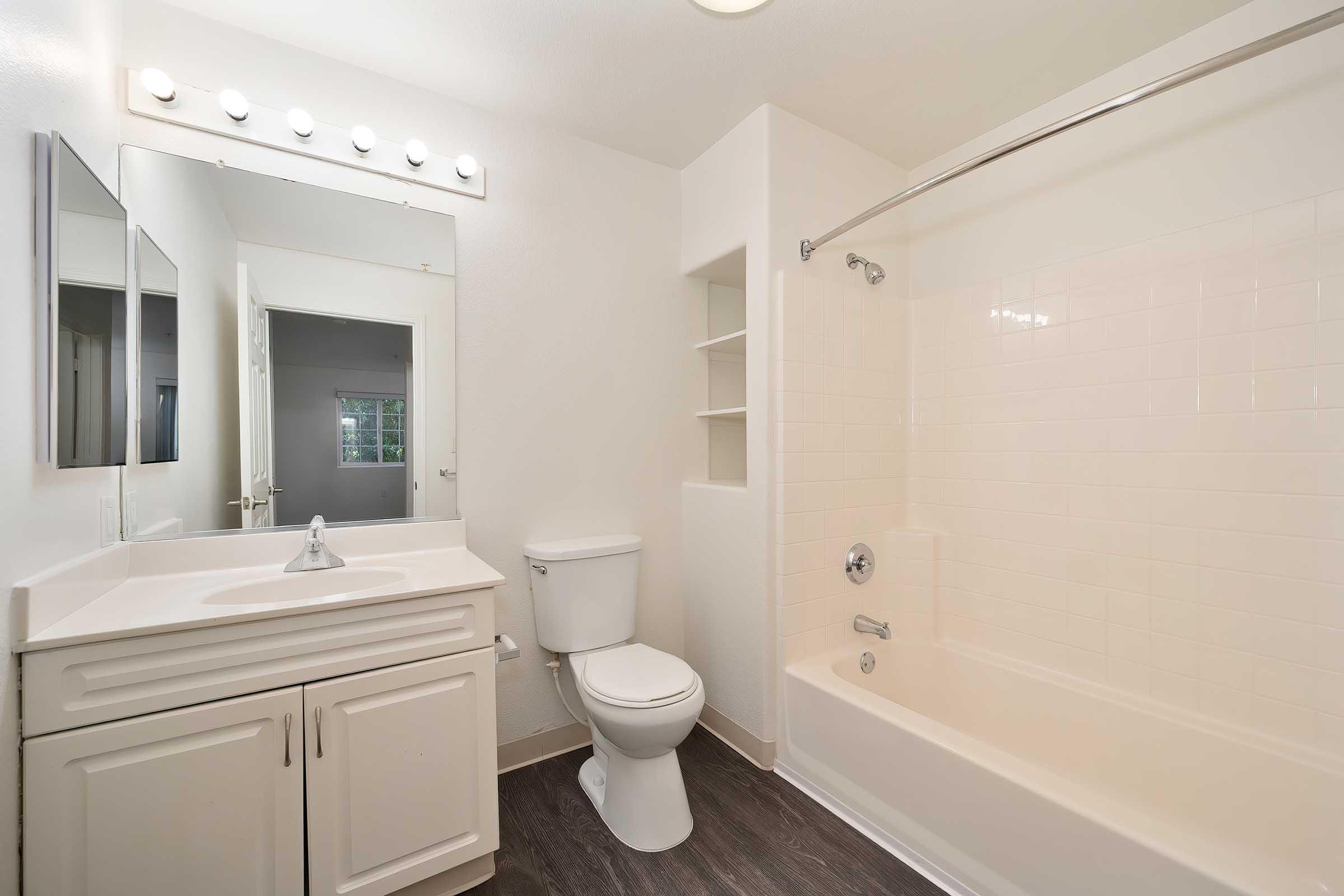 A clean, modern bathroom featuring a white bathtub with a shower, a single sink vanity with storage below, a toilet, and a large mirror illuminated by lights. There is a shelving unit next to the bathtub and a door in the background leading to another room. The walls are painted in a light color, enhancing the spacious feel.