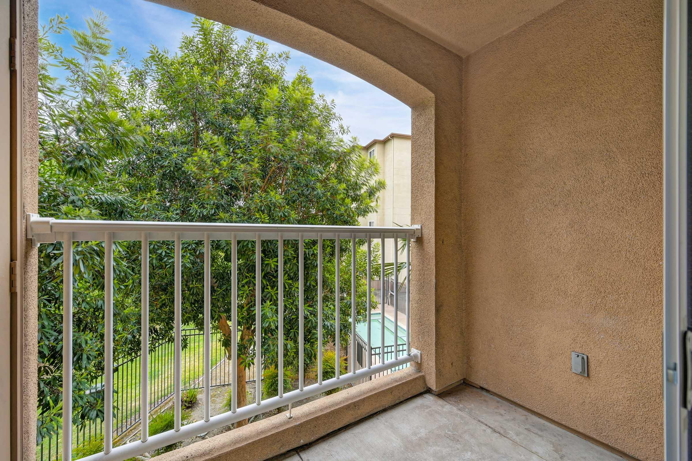 View from a balcony showcasing a light brown stucco wall and a white railing. A lush green tree is visible in the foreground, while an apartment complex can be seen in the background, hinting at a well-maintained outdoor space. The sky is partly cloudy, adding a serene ambiance to the scene.