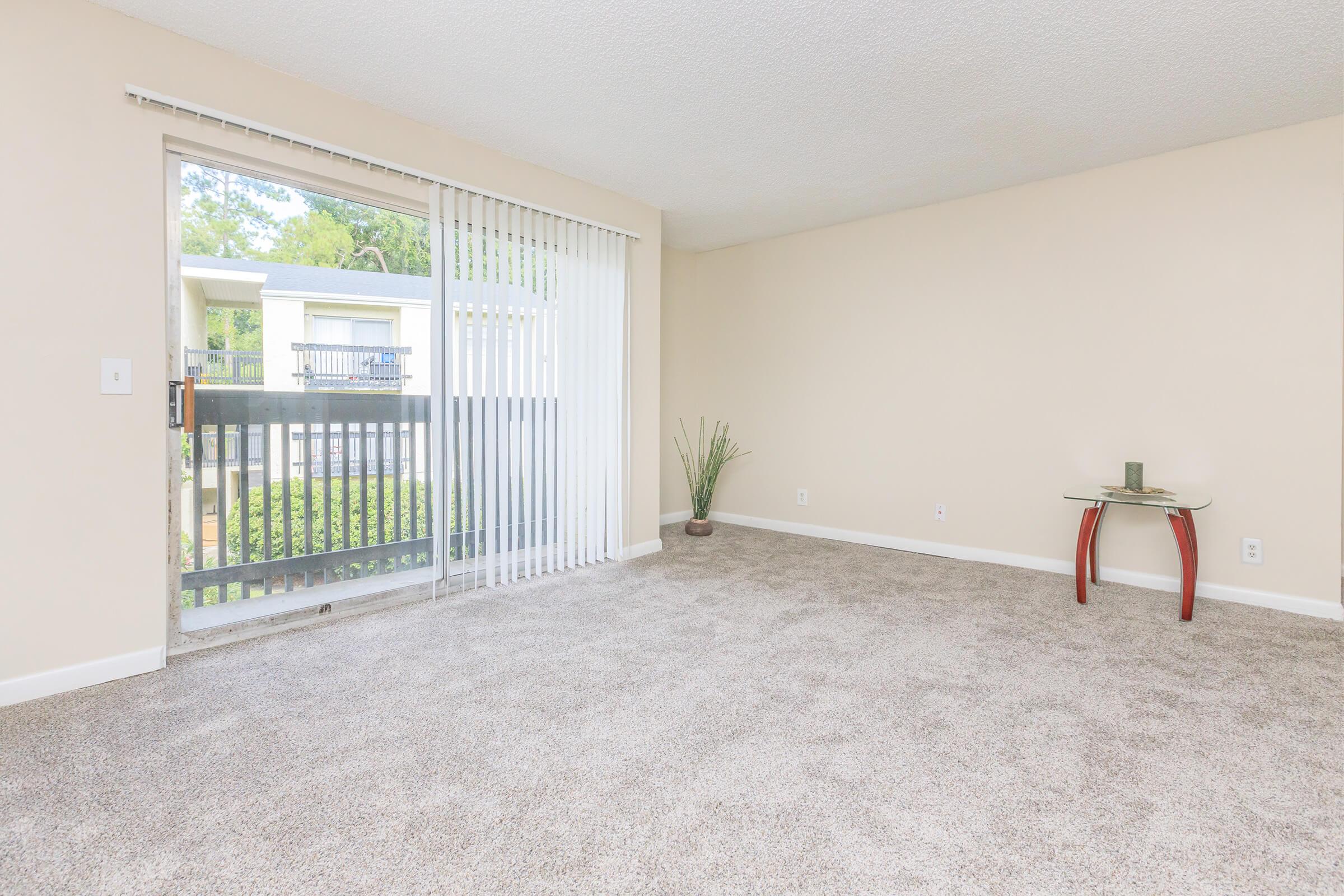 A spacious, empty living room featuring light beige walls and plush carpet. There is a large sliding glass door with vertical blinds leading to a balcony, allowing natural light to enter. A small glass table with curved legs is placed on the right side of the room, with a potted plant in the corner.