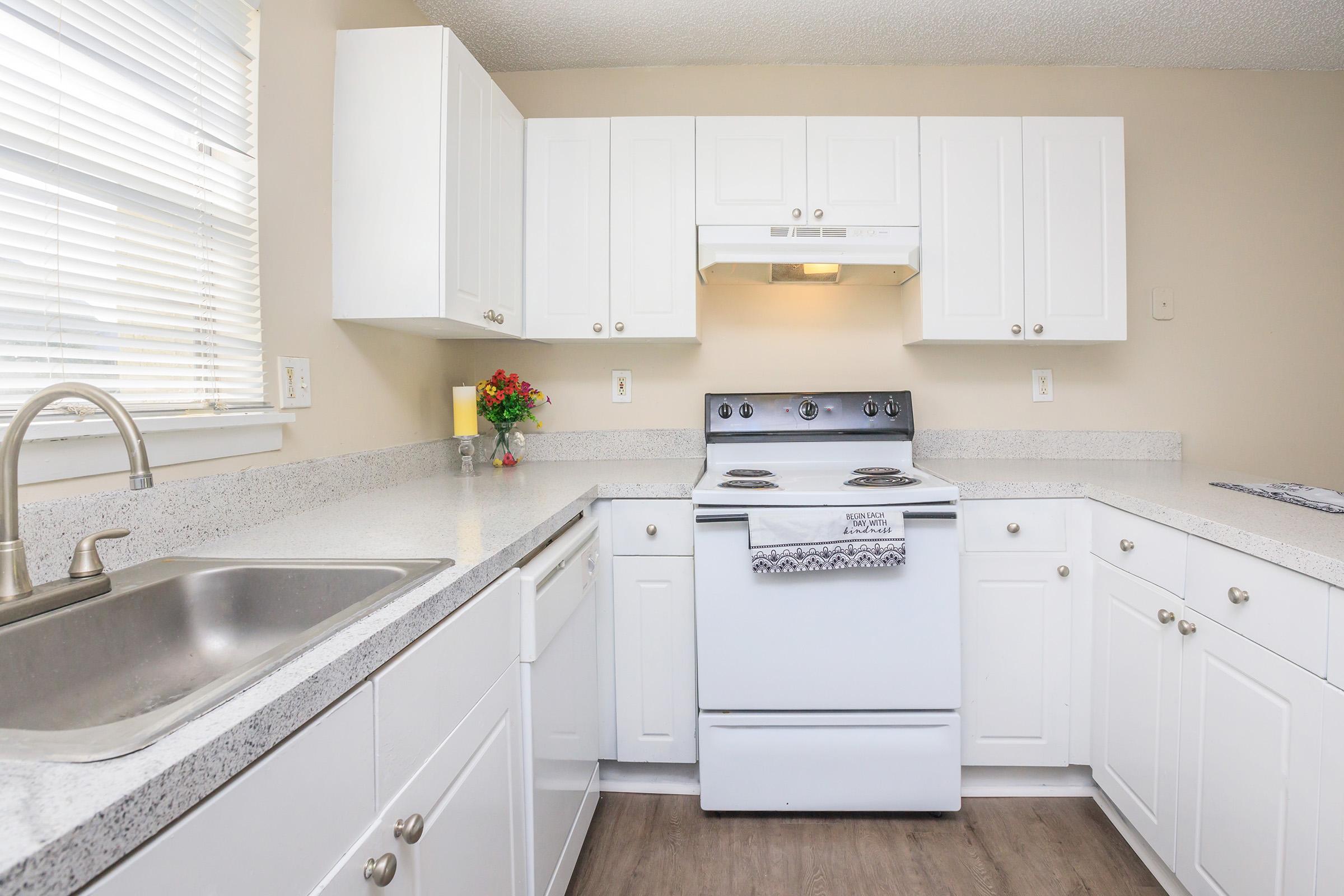 Modern kitchen featuring white cabinetry, a stainless steel sink, and a stove with an overhead vent. Countertops are light-colored, and there are artificial flowers on the counter. The space is well-lit with natural light coming from a nearby window. The flooring is a warm wood tone.