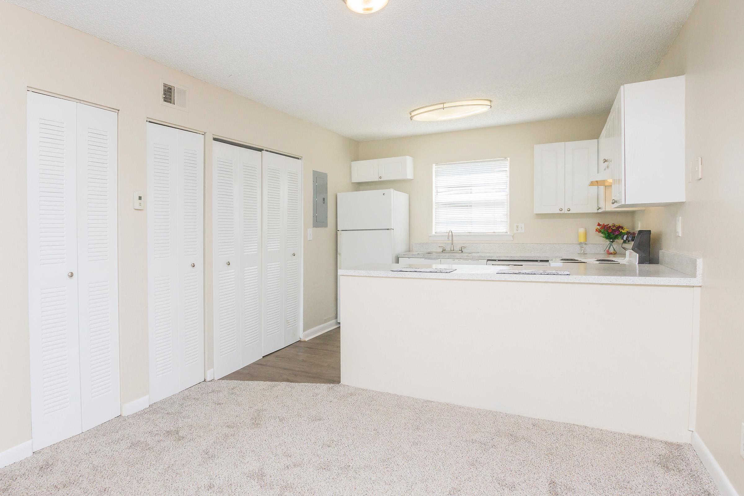 A bright, modern kitchen area with a small island, featuring white cabinets and a refrigerator. There are two closet doors on the left and a window providing natural light. The flooring appears to be light-colored carpet, creating a cozy atmosphere. Fresh flowers are visible on the counter.