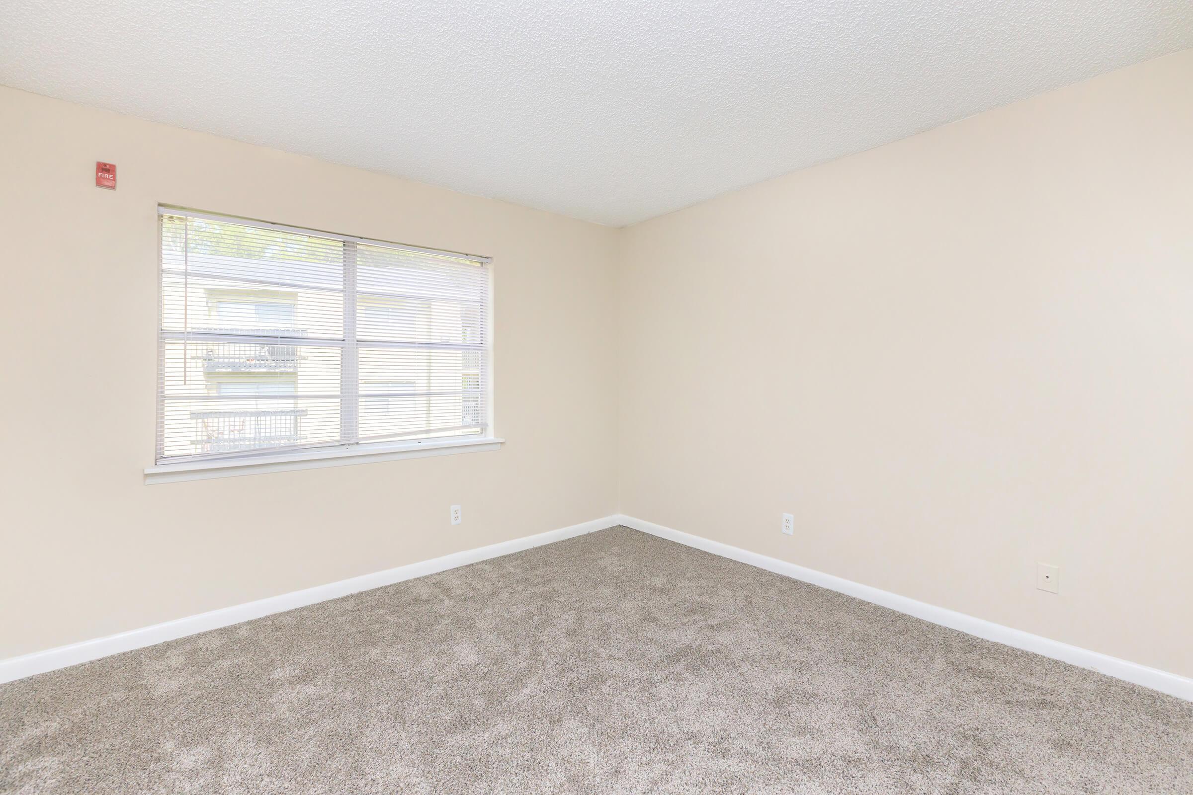 Empty room with light beige walls and beige carpet. A large window with horizontal blinds allows natural light to fill the space. The room has minimal furnishings and features a smooth ceiling. It appears spacious and is ready for personal decoration.