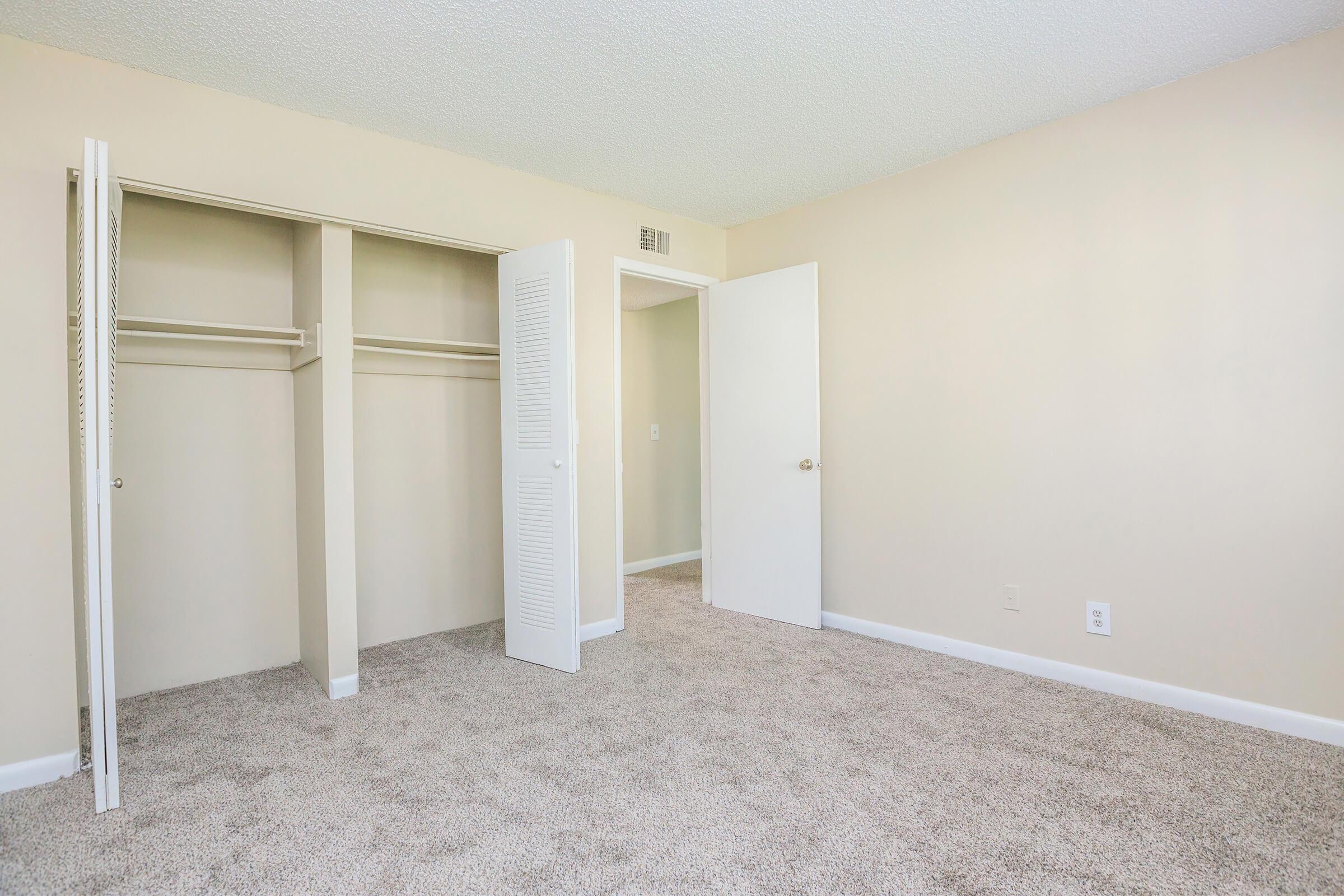A clean and empty bedroom featuring beige walls, a plush carpeted floor, and a closet with sliding doors. A white door leads to another room, enhancing the spacious feel of the area. The lighting is bright and inviting, creating a comfortable atmosphere.