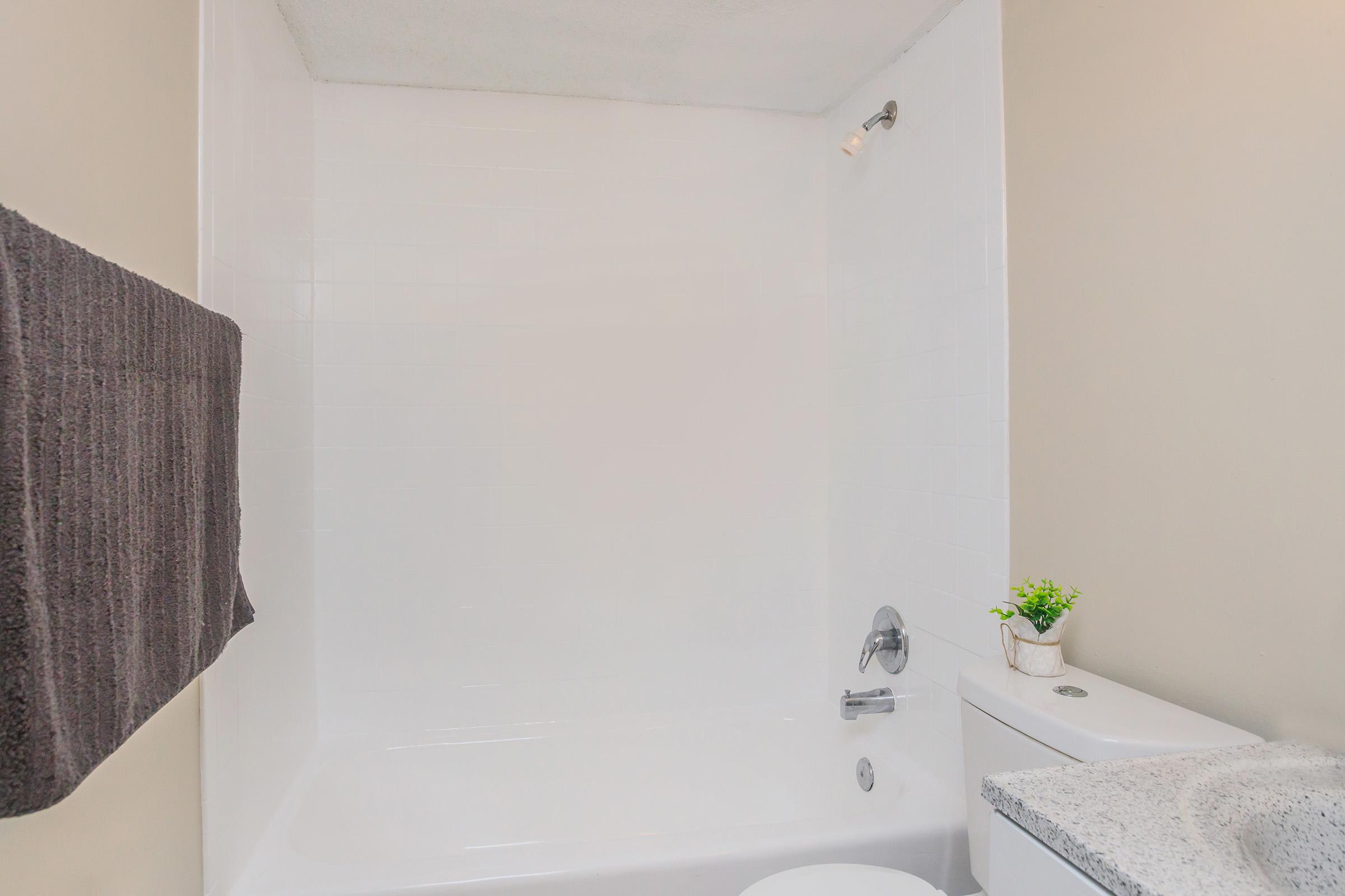A clean, modern bathroom featuring a white bathtub and shower with a light-colored wall. A gray towel hangs on the wall, and a small plant is placed on the countertop beside a white toilet. The overall design is minimalistic and bright, emphasizing simplicity and cleanliness.