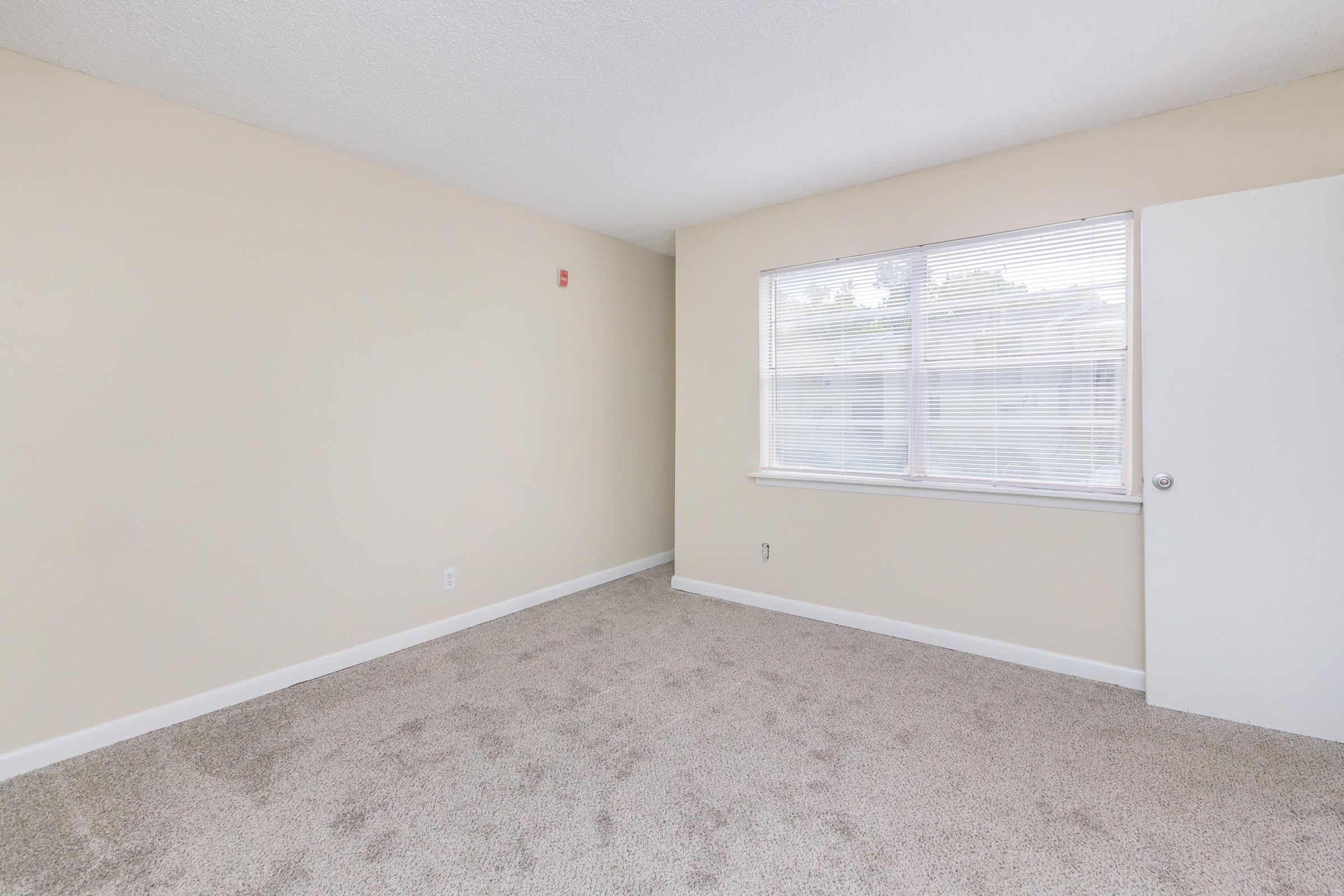 A spacious, vacant room with beige walls and light-colored carpet. A large window with white blinds lets in natural light, and there is a white door on the right side of the room. The overall appearance is clean and inviting, suitable for various uses.