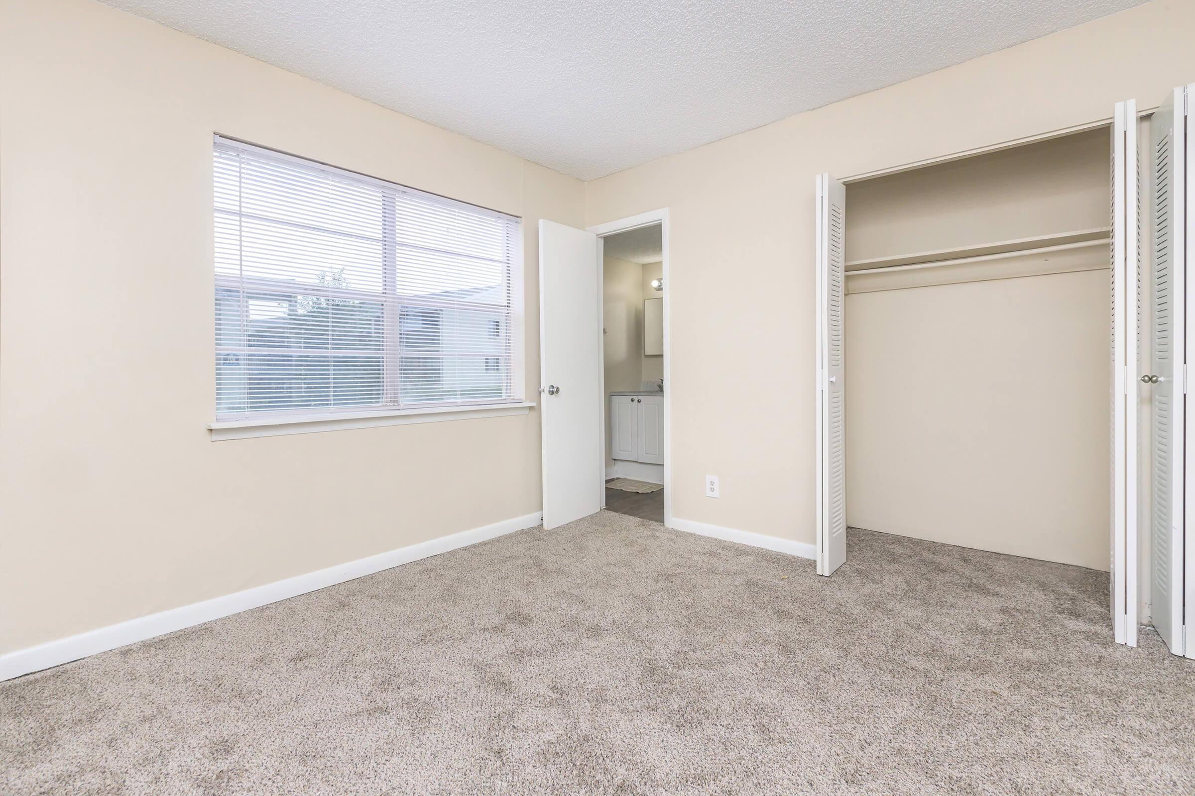 A light-colored room with beige walls and carpet. A large window lets in natural light, and there is a door leading to a bathroom visible in the background. A closet with sliding doors is on the right side. The room appears clean and spacious, suitable for various uses.