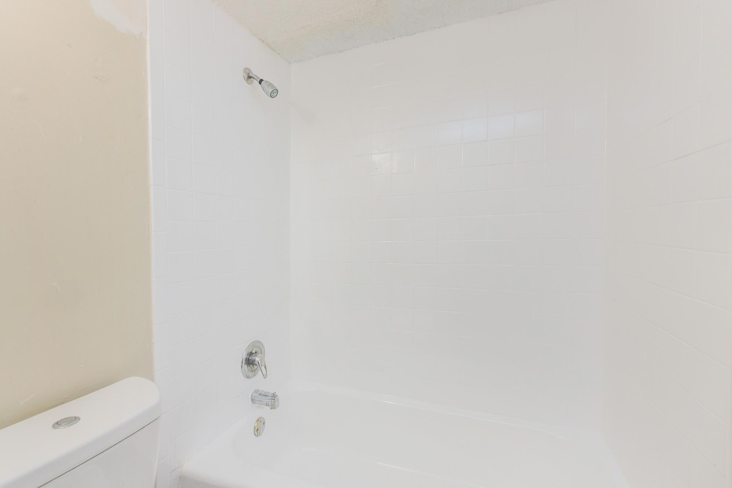 A clean and empty bathroom featuring a white tiled shower and bathtub. The walls are painted in a light color, and there is a shower fixture on the wall. A toilet is visible on the left side of the image. The overall appearance is bright and minimalist.