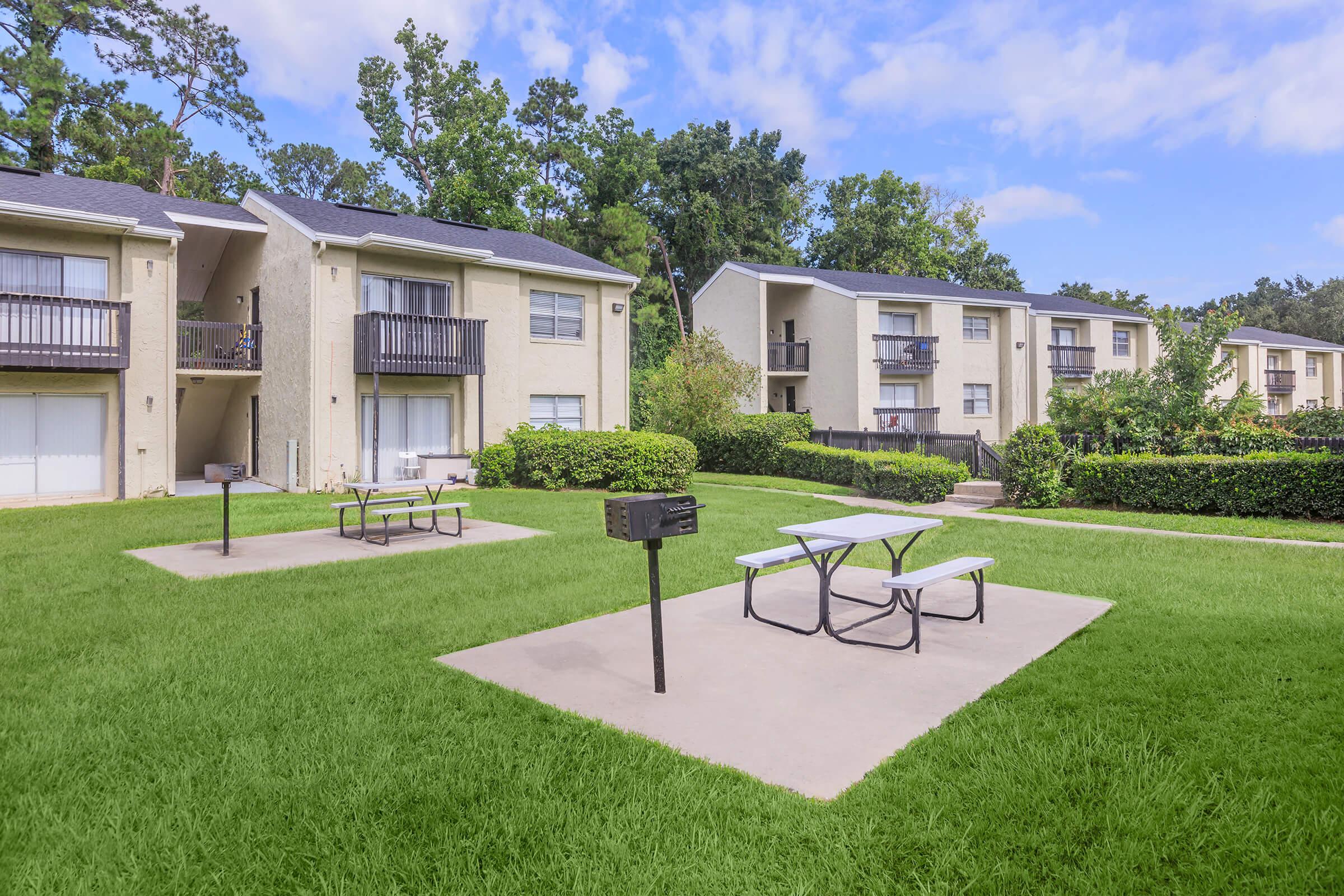 View of a grassy courtyard with picnic tables and a grill, surrounded by two apartment buildings. Lush trees and landscaping enhance the outdoor space, creating a welcoming area for residents to gather and enjoy the outdoors. The sky is partly cloudy, adding a pleasant atmosphere to the setting.