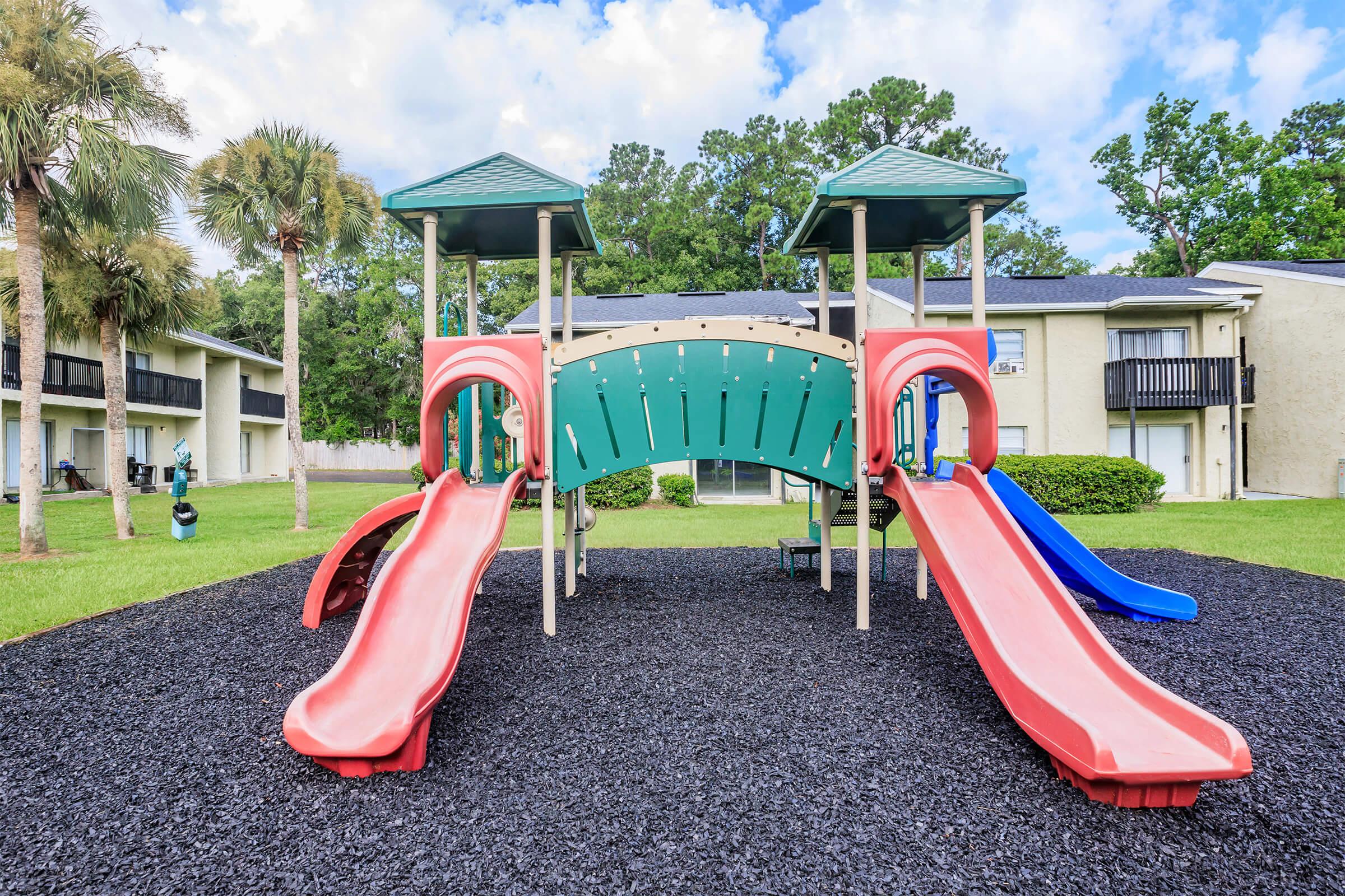 Colorful playground equipment featuring two slides, surrounded by black rubber mulch and palm trees, set against a backdrop of residential buildings and a blue sky with clouds.