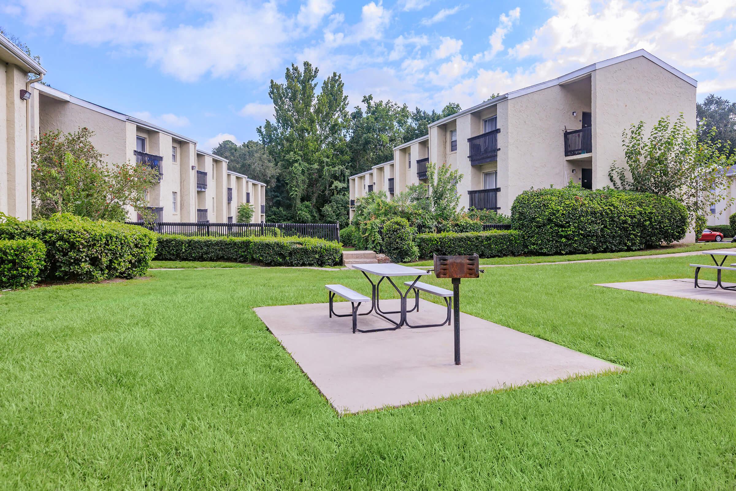 A grassy courtyard with two picnic tables on concrete slabs, surrounded by shrubs and trees. In the background, there are three multi-story apartment buildings with balconies under a blue sky with scattered clouds.