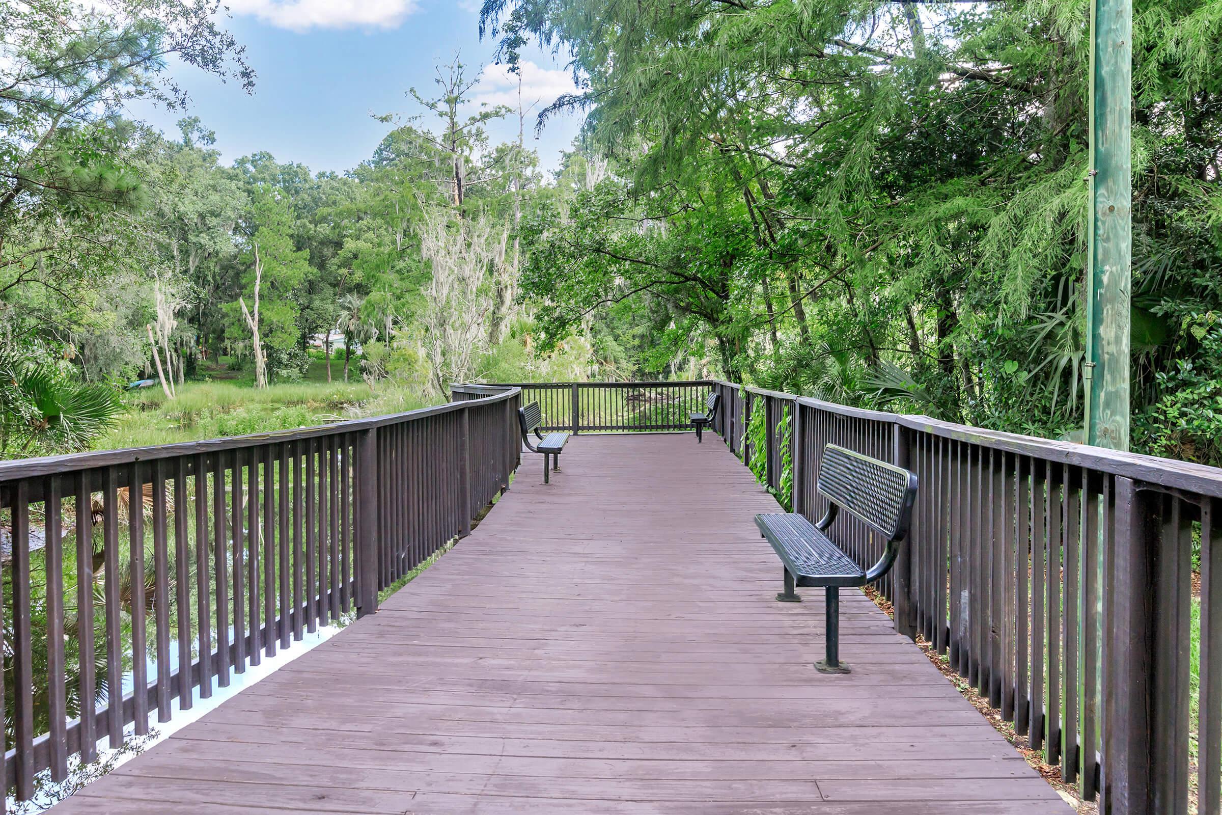 A wooden walkway with black metal benches on either side, surrounded by lush greenery and trees. The path extends into a serene natural area with water visible along the edges, under a clear blue sky.