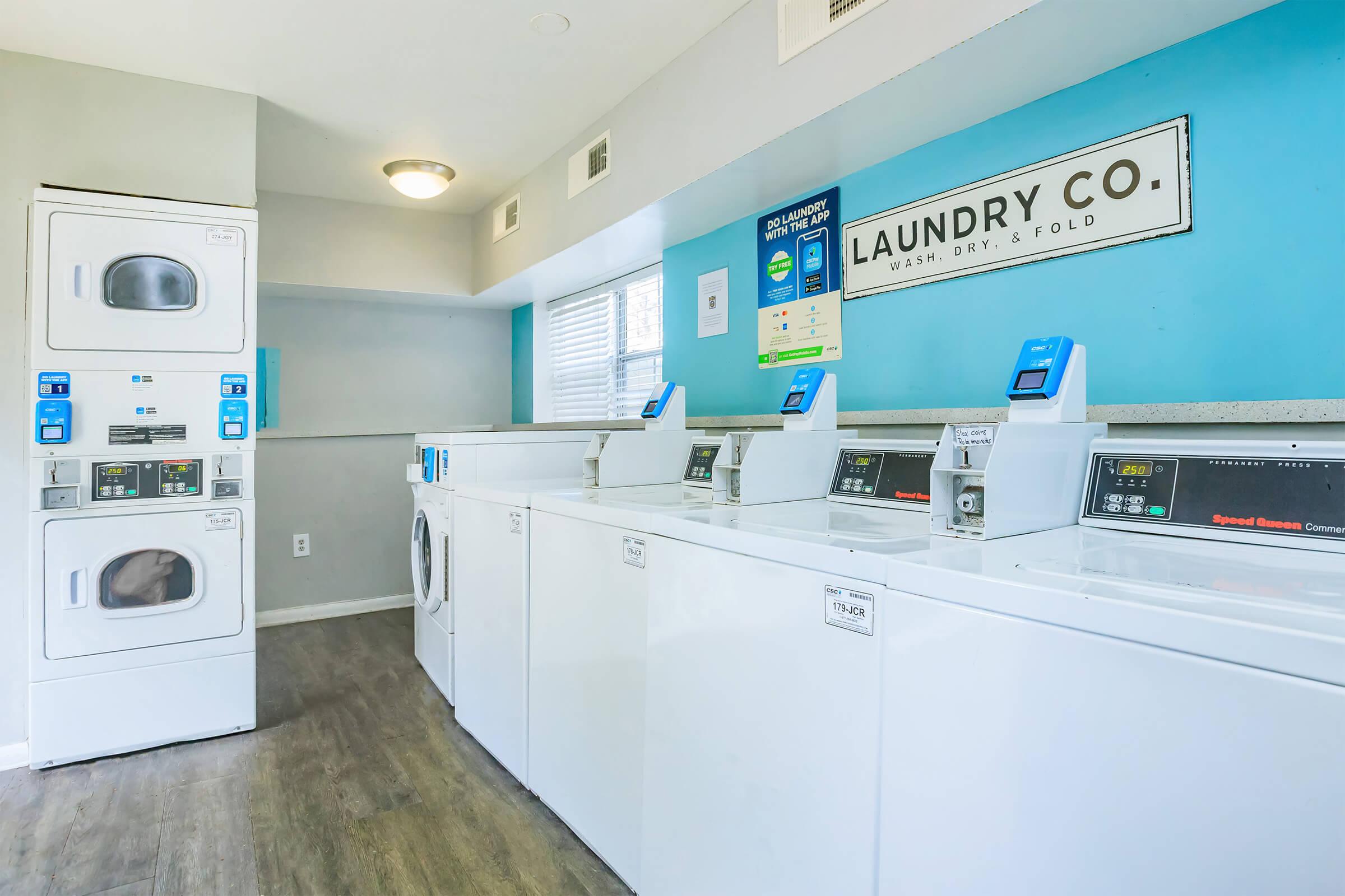 A clean and modern laundry room featuring several white washing machines and dryers. There's a large sign that says "LAUNDRY CO." on the wall, and the space is well-lit with windows. The machines have digital displays indicating their features. The flooring is a light wood, enhancing the bright atmosphere.