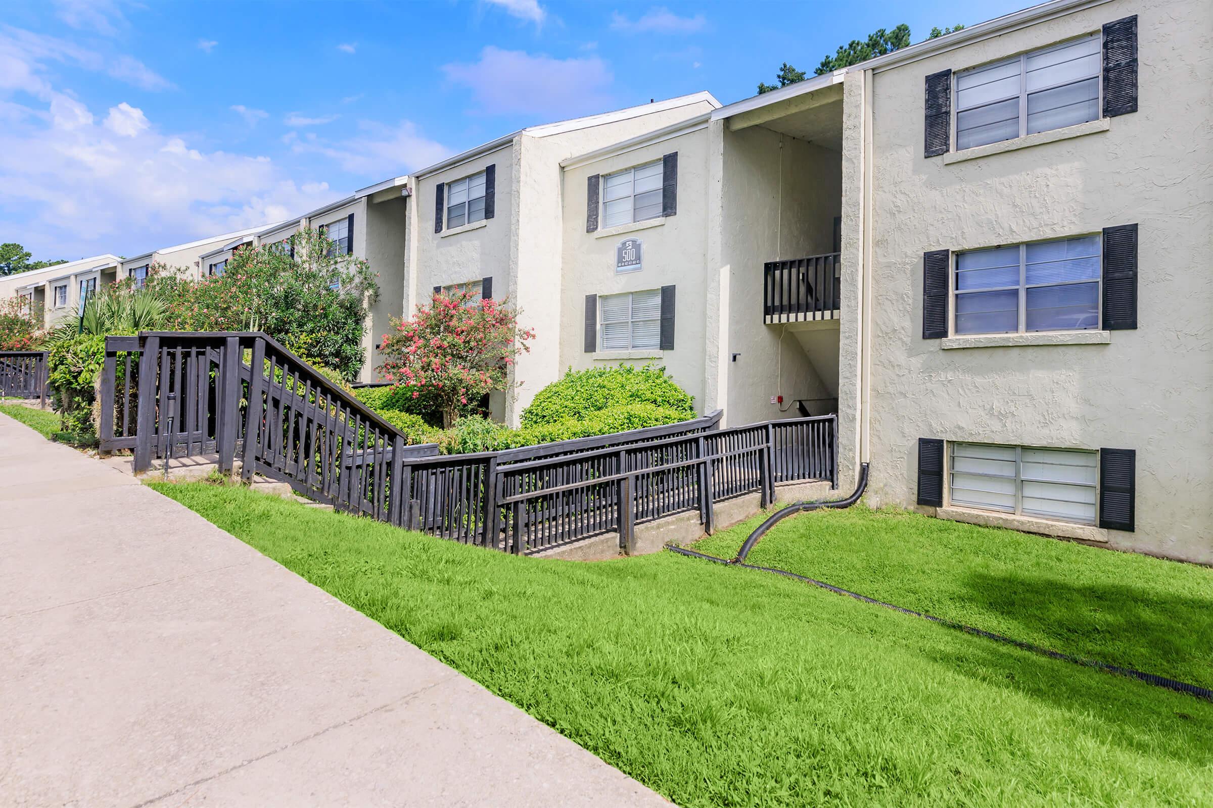 Exterior view of a multi-unit apartment building with a light-colored facade. The structure features black window shutters and is surrounded by neatly trimmed grass and vibrant foliage. A sloped walkway with a wooden railing leads up to the entrance, under a partly cloudy sky.