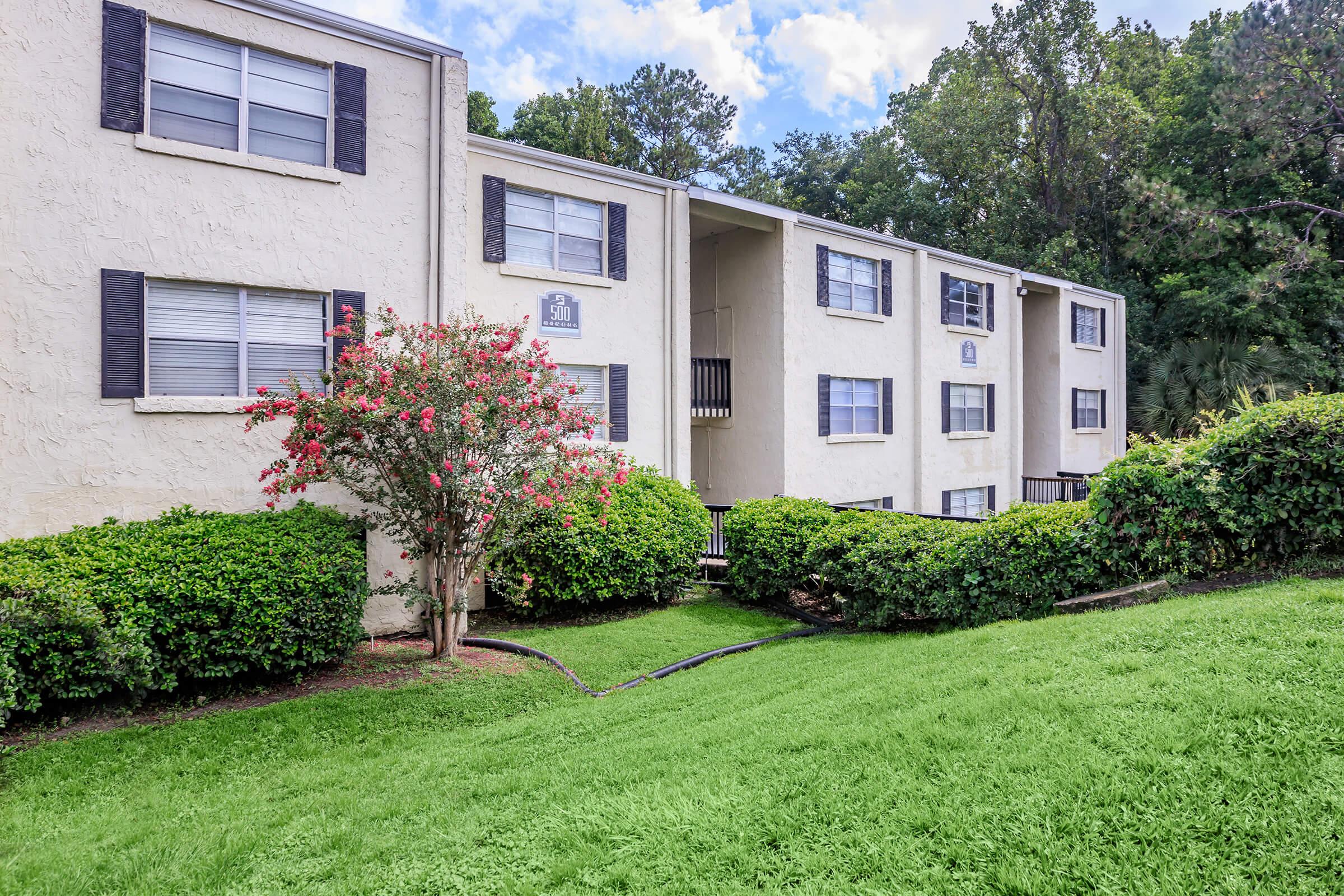 A two-story residential building with light-colored walls and dark shutters. In front, there is a well-maintained lawn with green grass and shrubs, including flowering plants. The scene is set against a backdrop of trees under a partly cloudy sky.