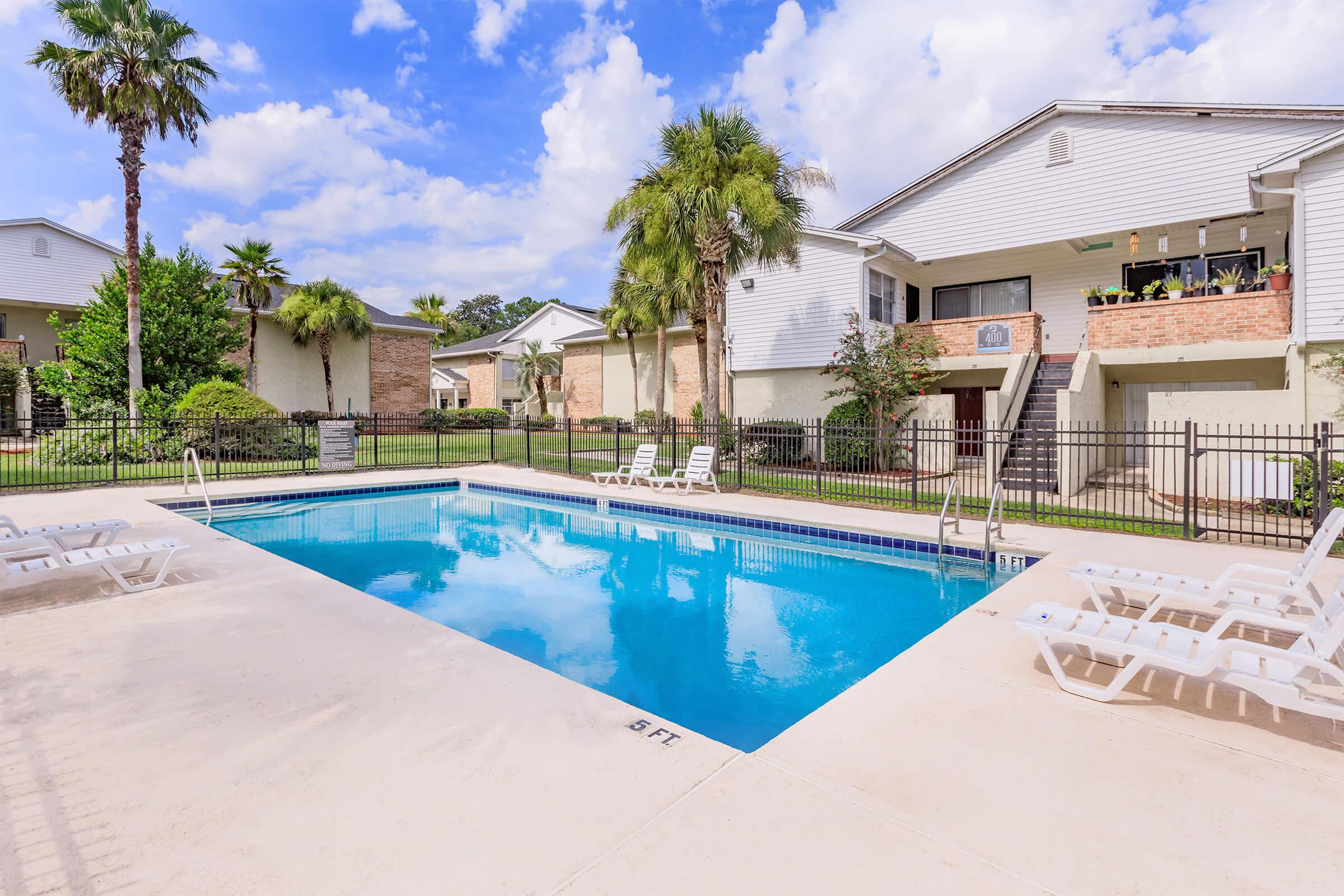 A clear swimming pool surrounded by lounge chairs, situated in a residential area with palm trees and greenery. Two buildings are visible in the background, featuring balconies and staircases. The sky is partly cloudy, creating a bright, inviting atmosphere.