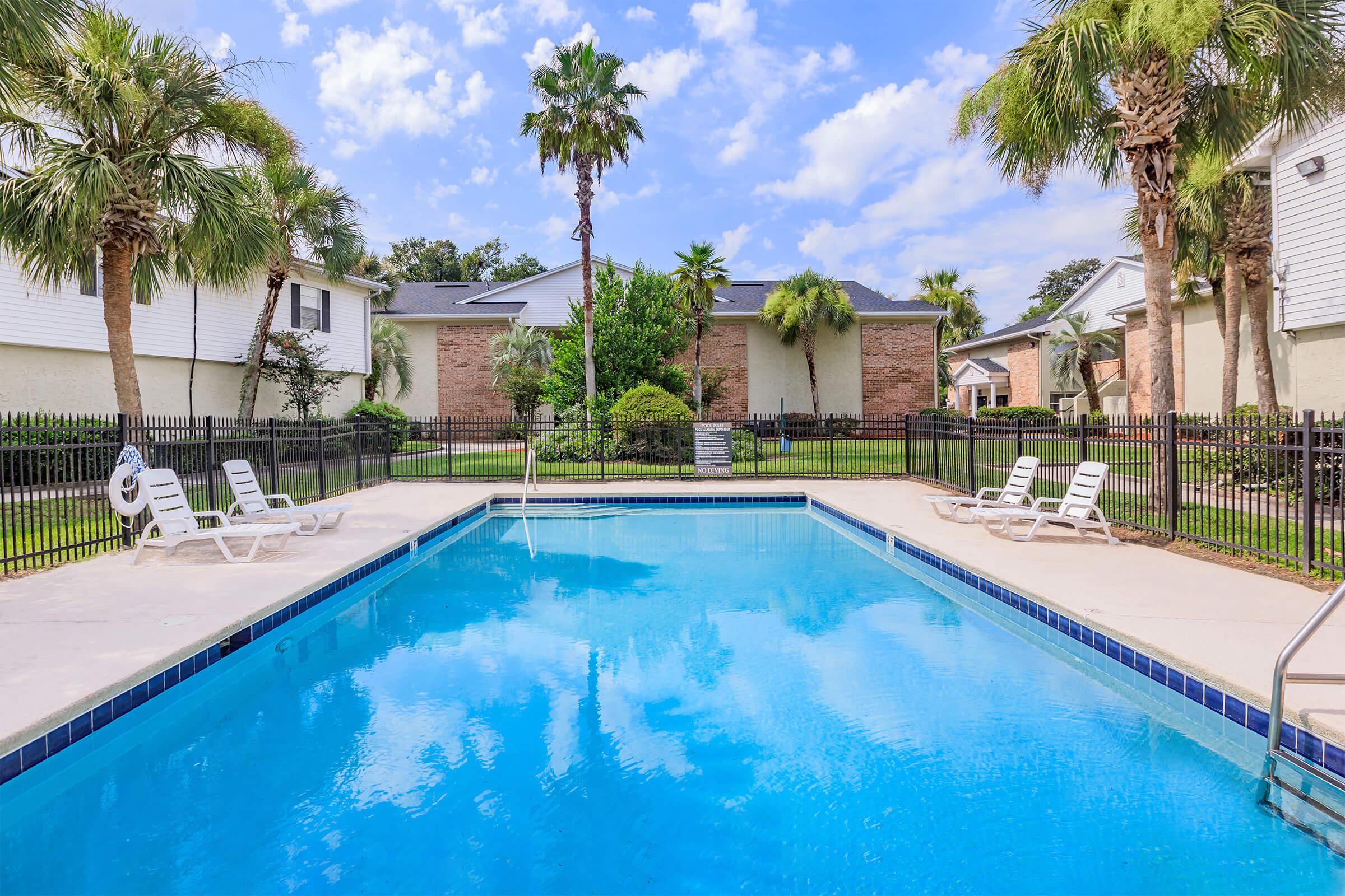 A clear blue swimming pool surrounded by lounge chairs and palm trees. The pool area features a fenced perimeter and views of landscaped gardens and buildings in the background under a bright, partly cloudy sky.